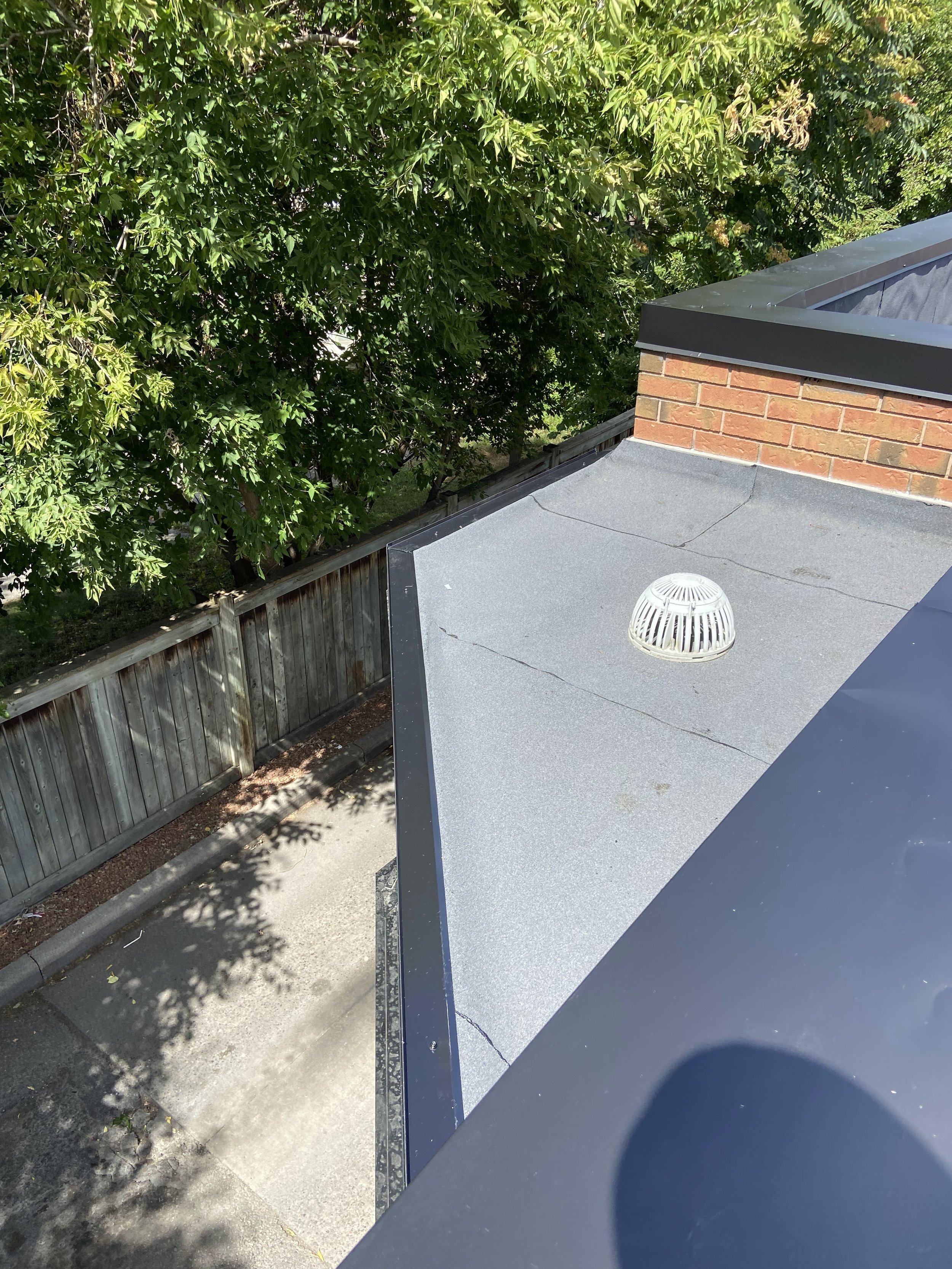 The image shows the corner of a flat rooftop with a grey surface, a red brick chimney, and a part of a metal railing. There is a tree with green leaves and branches behind a wooden fence, with a shadow cast on the ground.