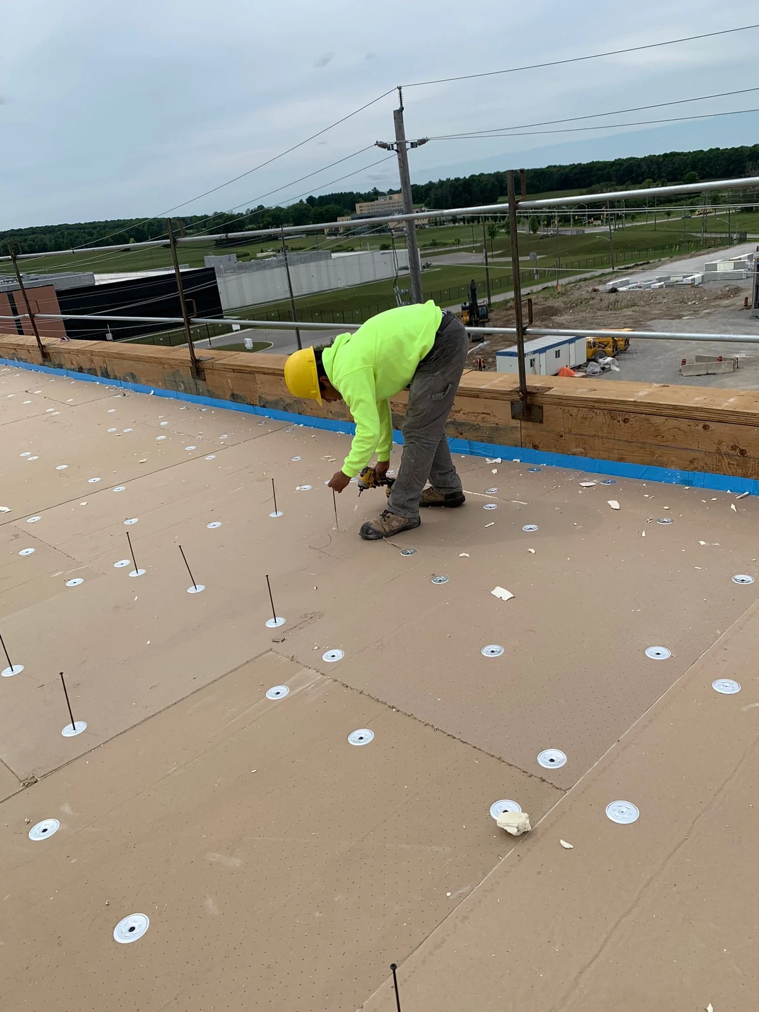 Construction worker wearing a yellow safety helmet and bright yellow shirt bent over working on a rooftop. The rooftop is under construction with circular fastener points and nails visible. In the background, there is a large green field, trees, buildings, and a cloudy sky.