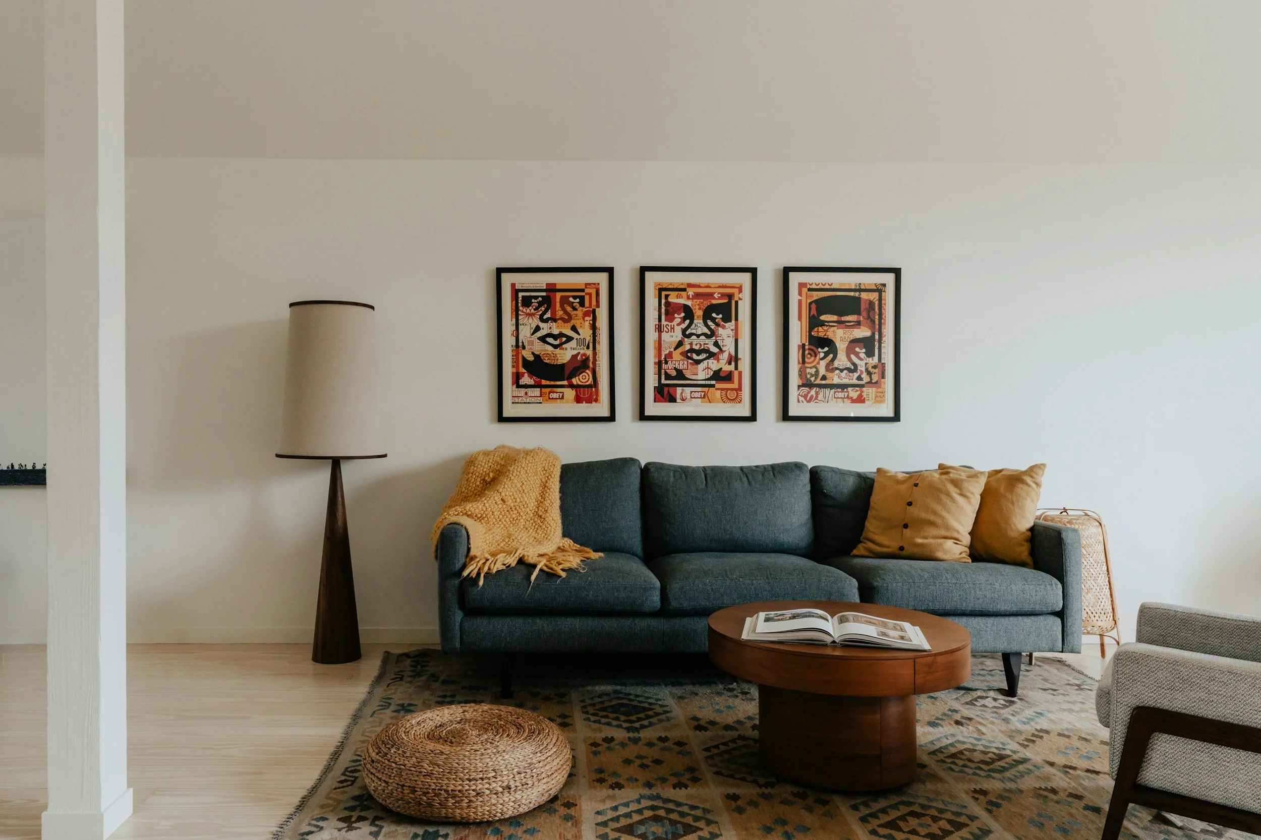Living room with a gray sofa, three framed abstract artworks on the wall, a tall lampshade on a wooden stand, a round woven floor cushion, a wooden coffee table with an opened magazine, a patterned area rug, and a beige armchair.