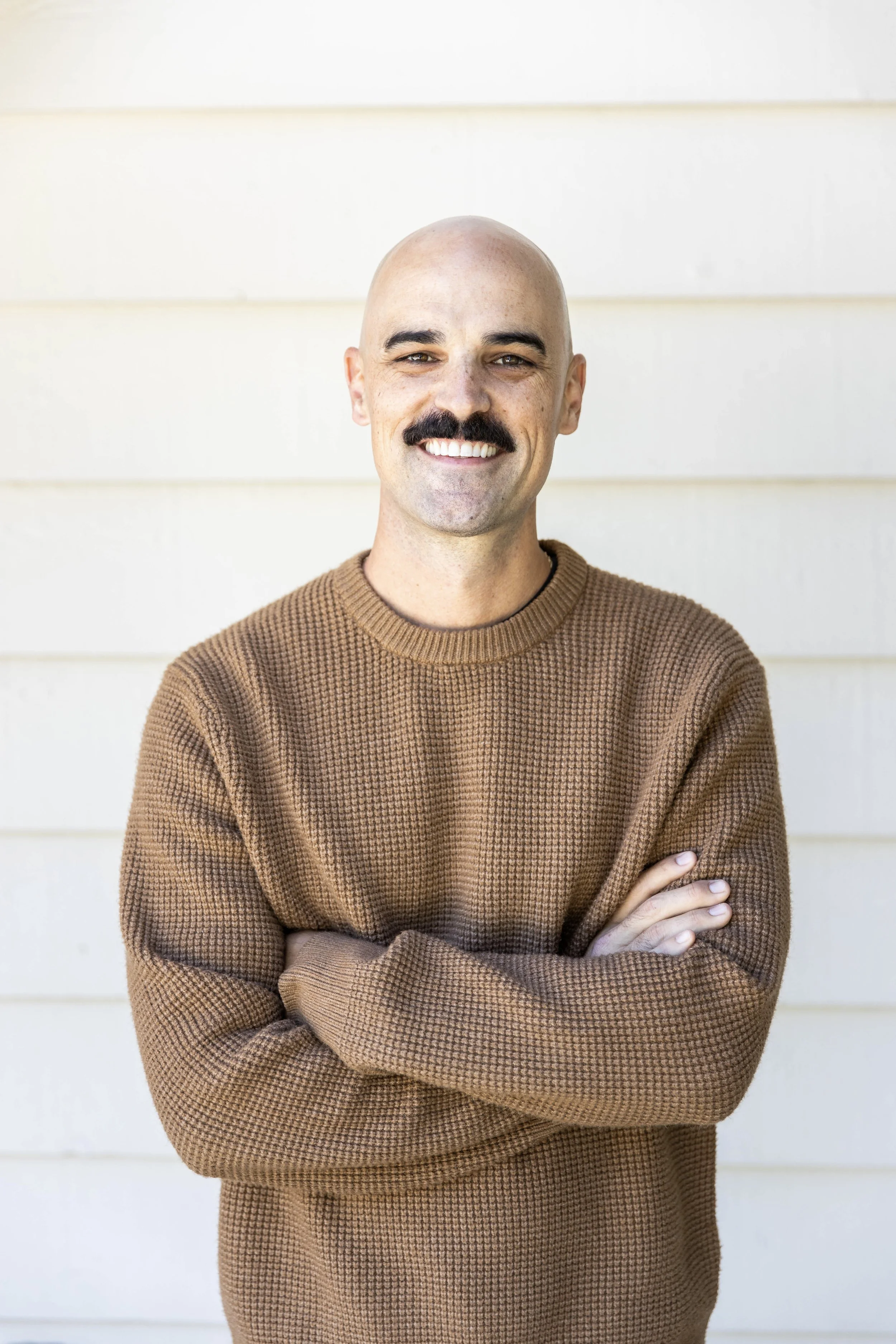 A man with a mustache and bald head is smiling and standing with arms crossed in front of a white-paneled wall, wearing a brown knitted sweater.