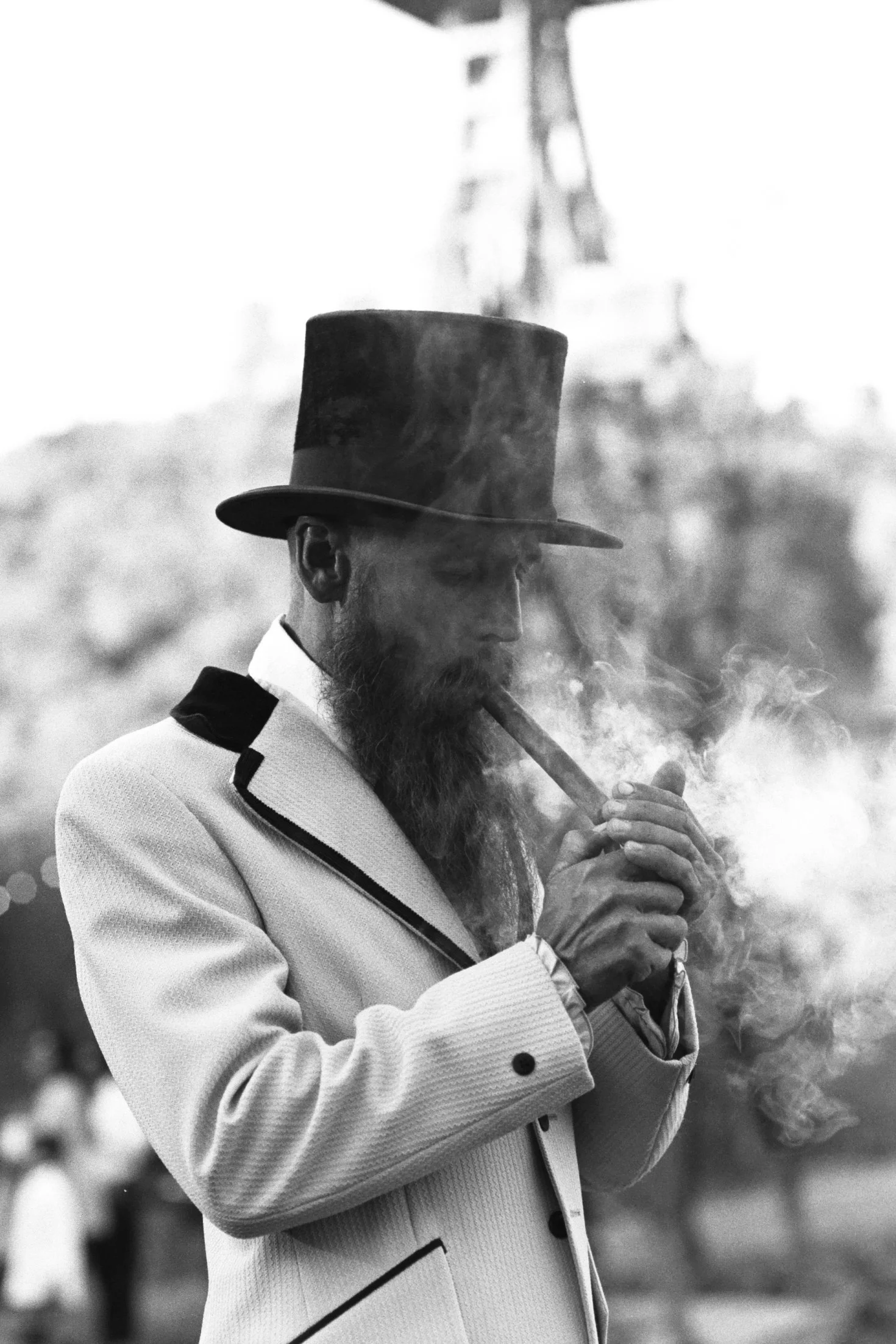 A man with a beard wearing a top hat and a light-colored jacket, smoking a pipe, is outdoors with a blurred background.