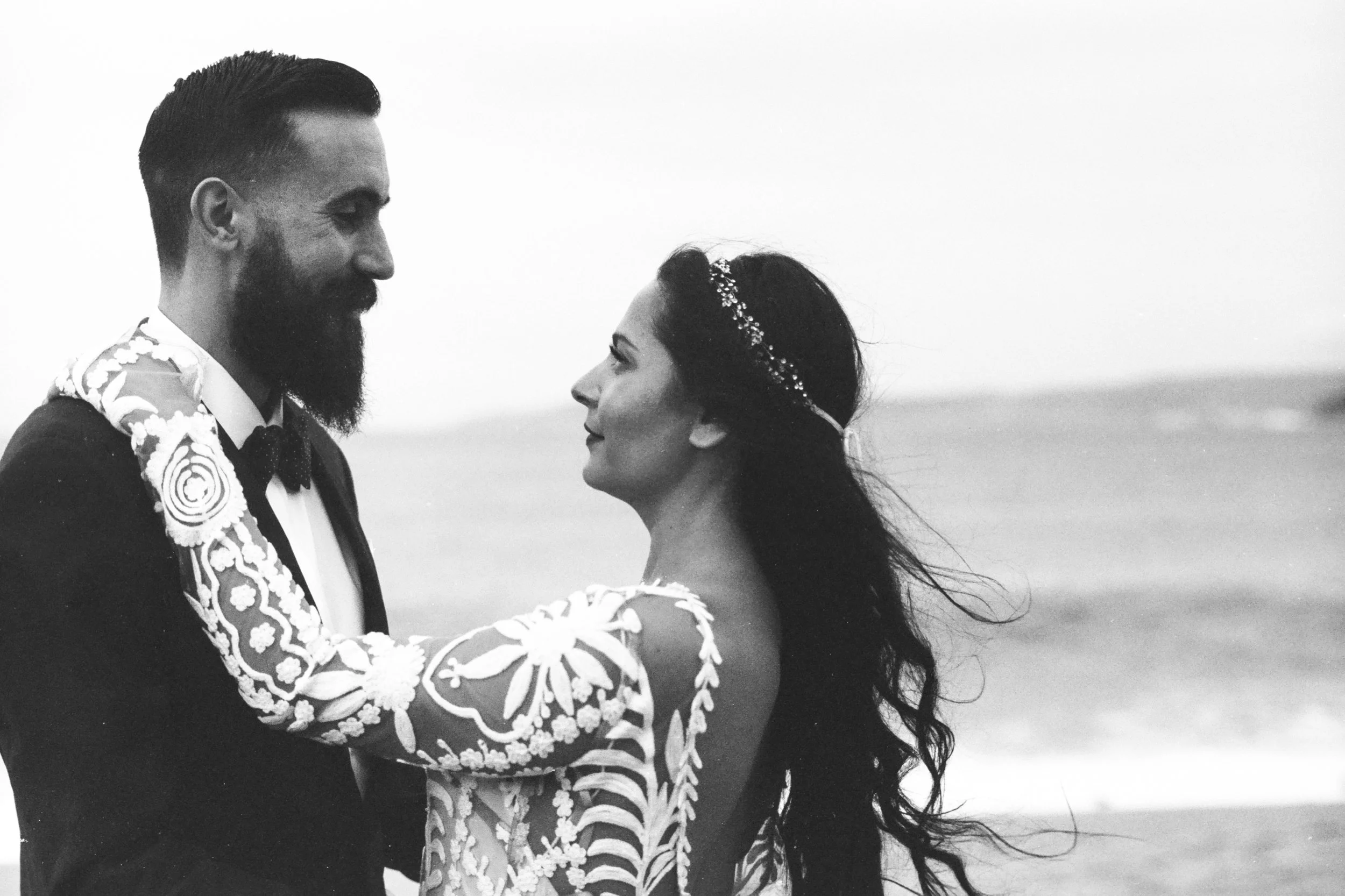 A black and white photo of a couple looking into each other's eyes. The man has a beard and is wearing a tuxedo with a floral boutonniere. The woman has long, dark hair with a decorative headband and is wearing a dress with floral embroidery. They ar