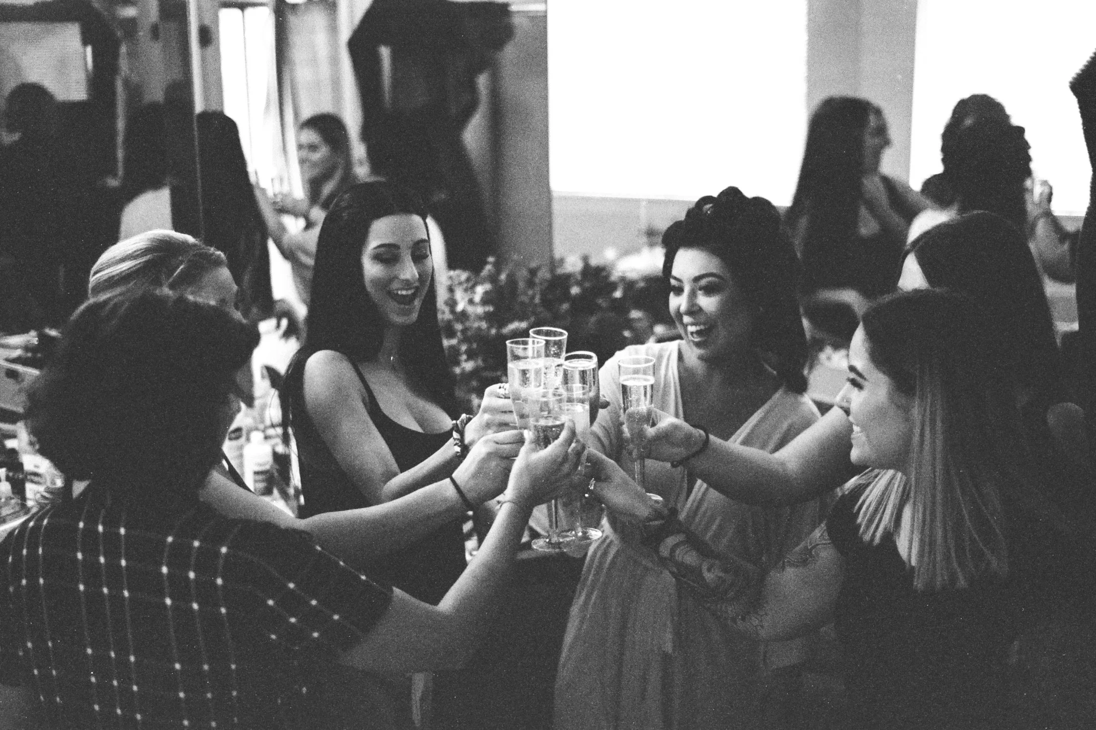 A black and white photo of a group of women gathered around a table, raising glasses in a toast and smiling at each other at a social event or party.