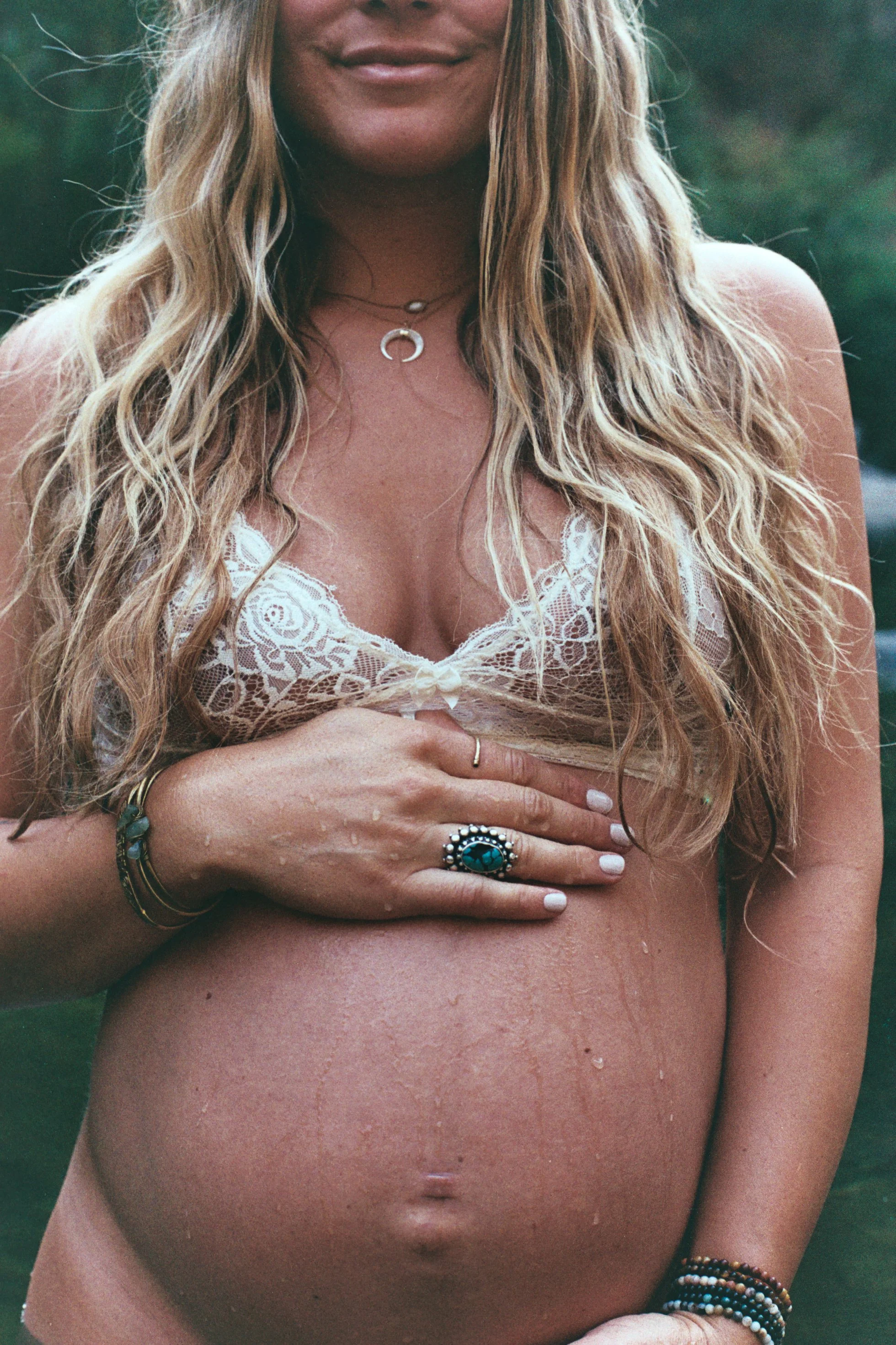 Close-up of a woman with long wavy blonde hair wearing jewelry and a lace bra, with her hand placed on her abdomen, outdoors with greenery in the background.
