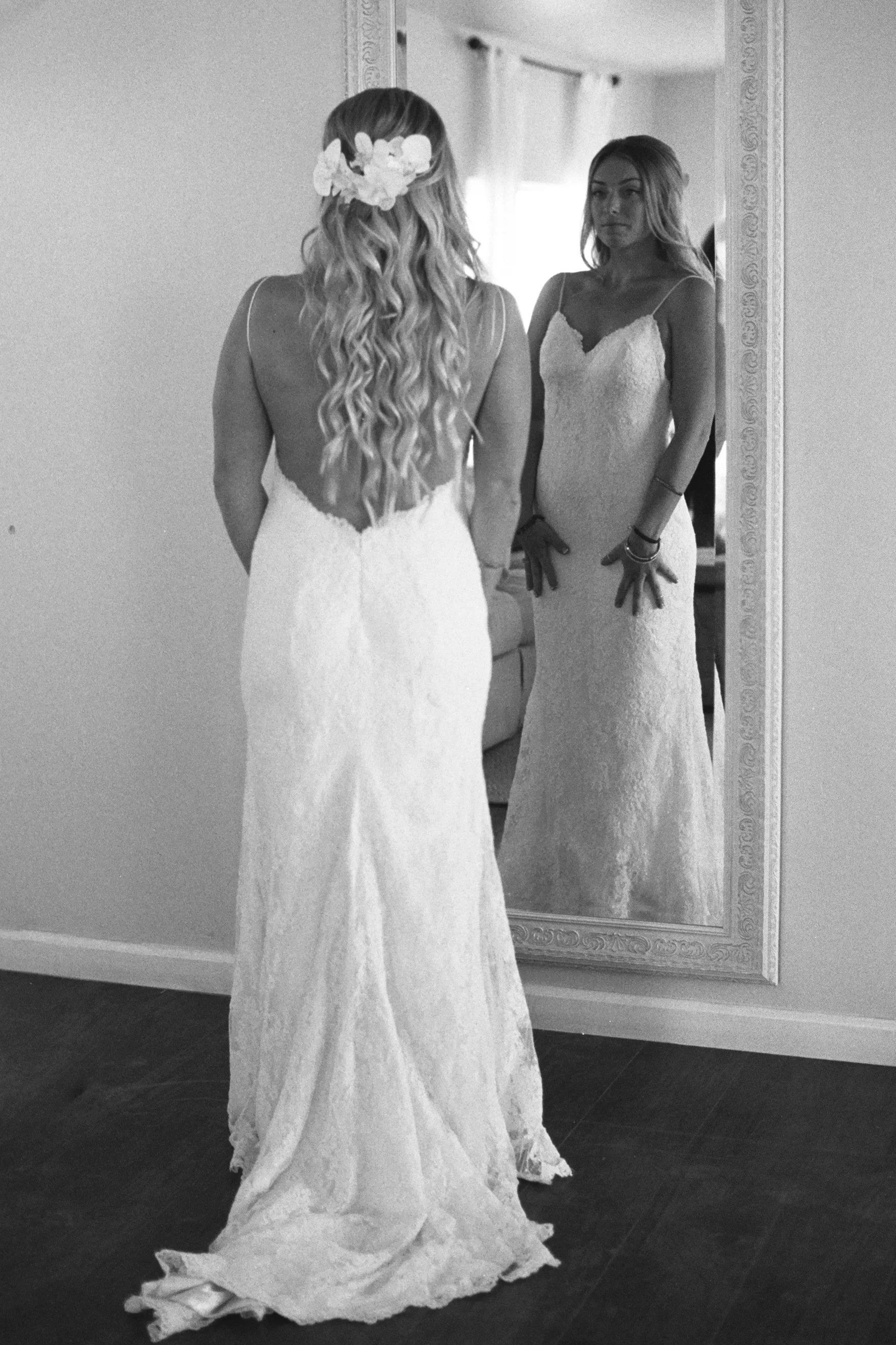 A woman in a wedding dress looking at herself in a large mirror. The dress has a lace design with a long train. The woman's hair is styled with loose waves and a floral hairpiece. The room has white walls and a window in the background.