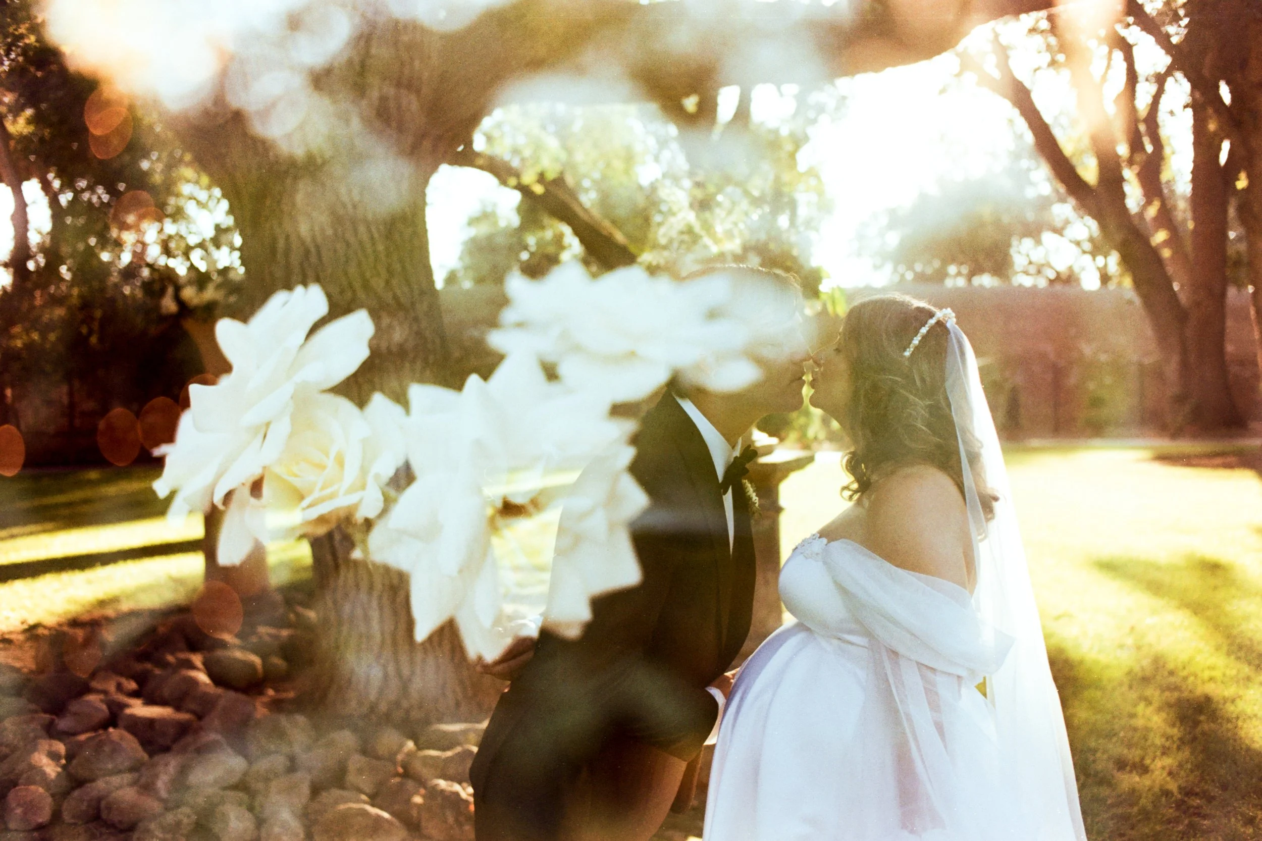 A bride and groom embrace outdoors during a wedding, with sunlight filtering through trees in the background. The photo has a dreamy, overexposed effect.