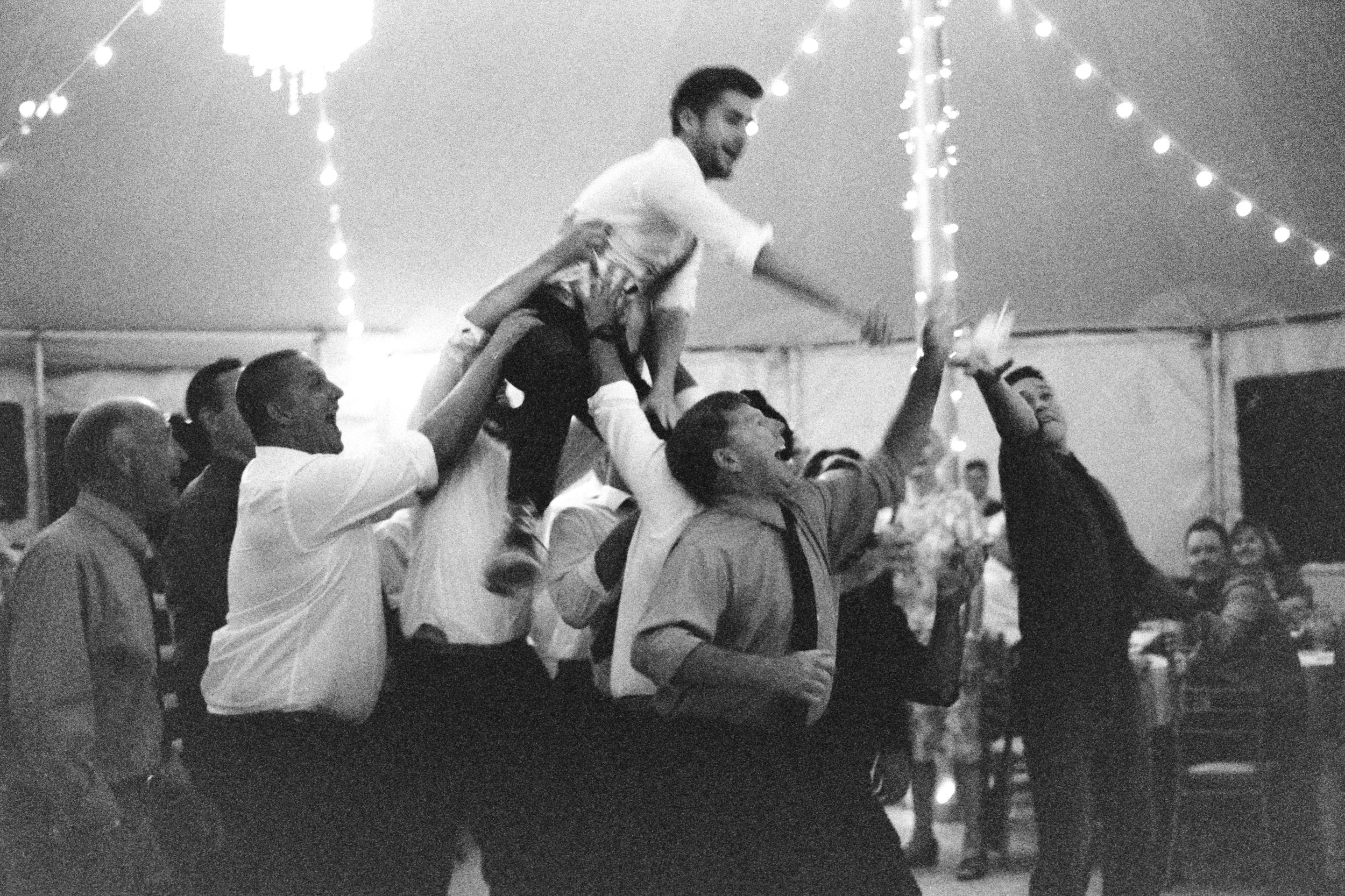 People celebrating at an event, with one person being lifted on others' shoulders, under string lights hanging from a tent ceiling.