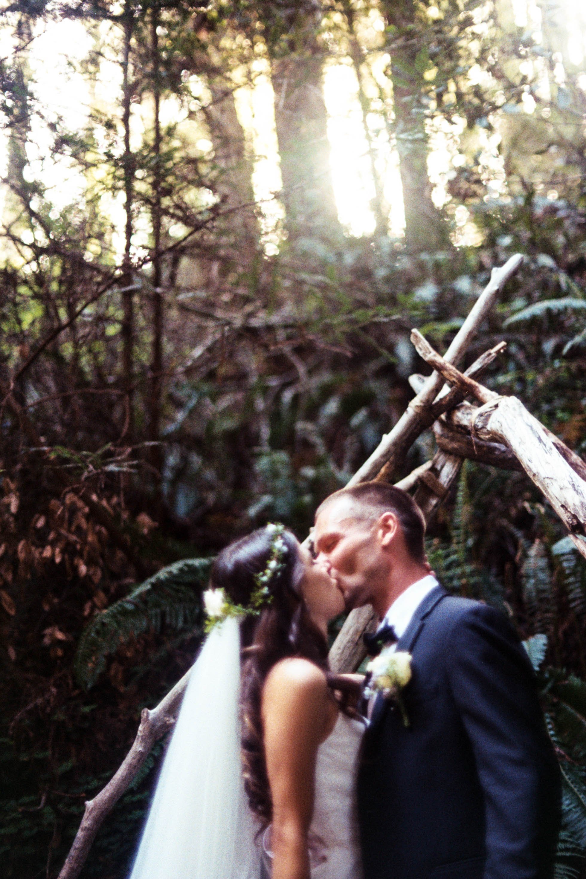 A bride and groom share a kiss during their wedding ceremony in a lush forest setting, with sunlight filtering through the trees.