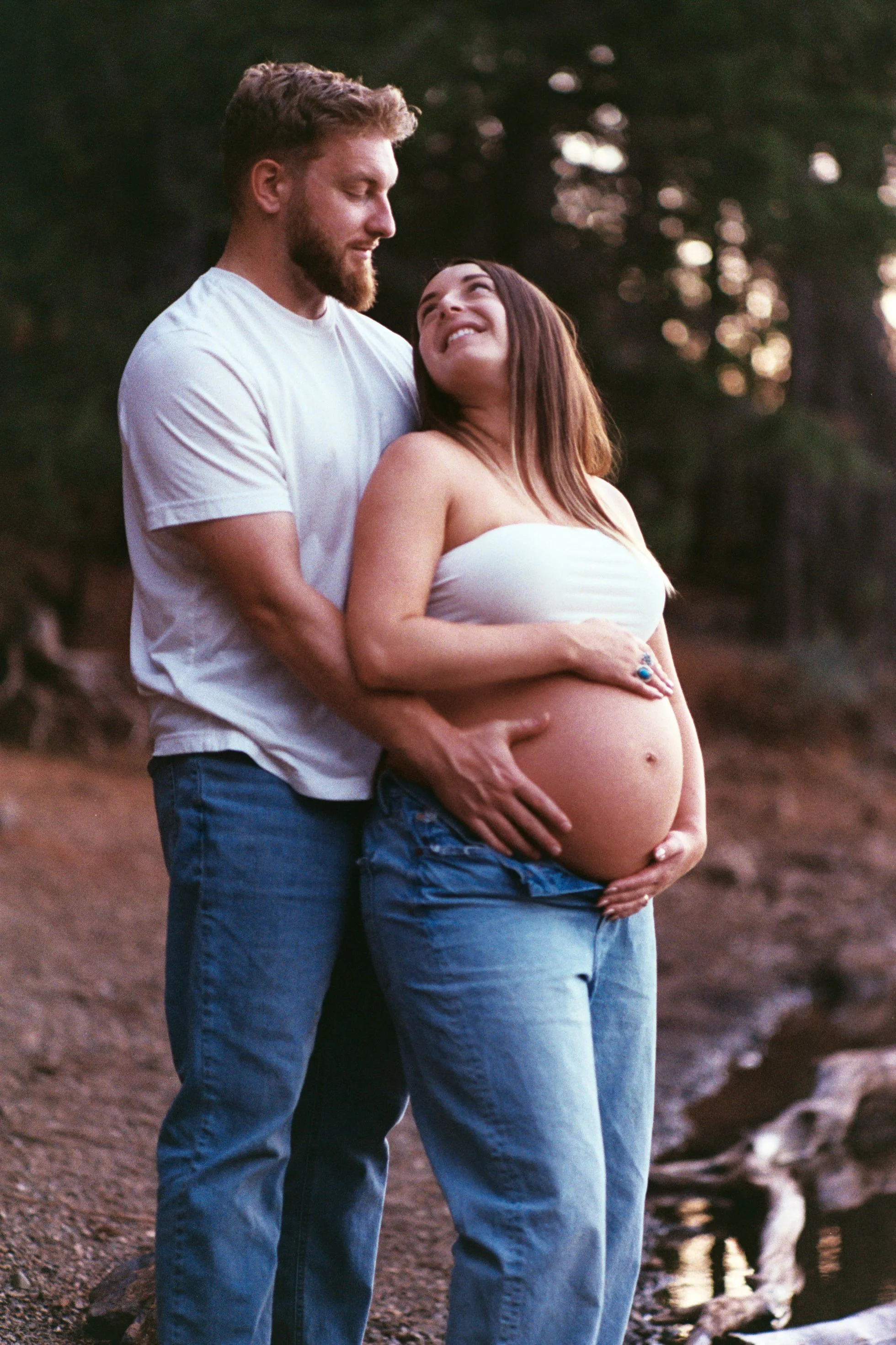 A pregnant woman and a man are standing outdoors, embracing each other and smiling warmly at each other.