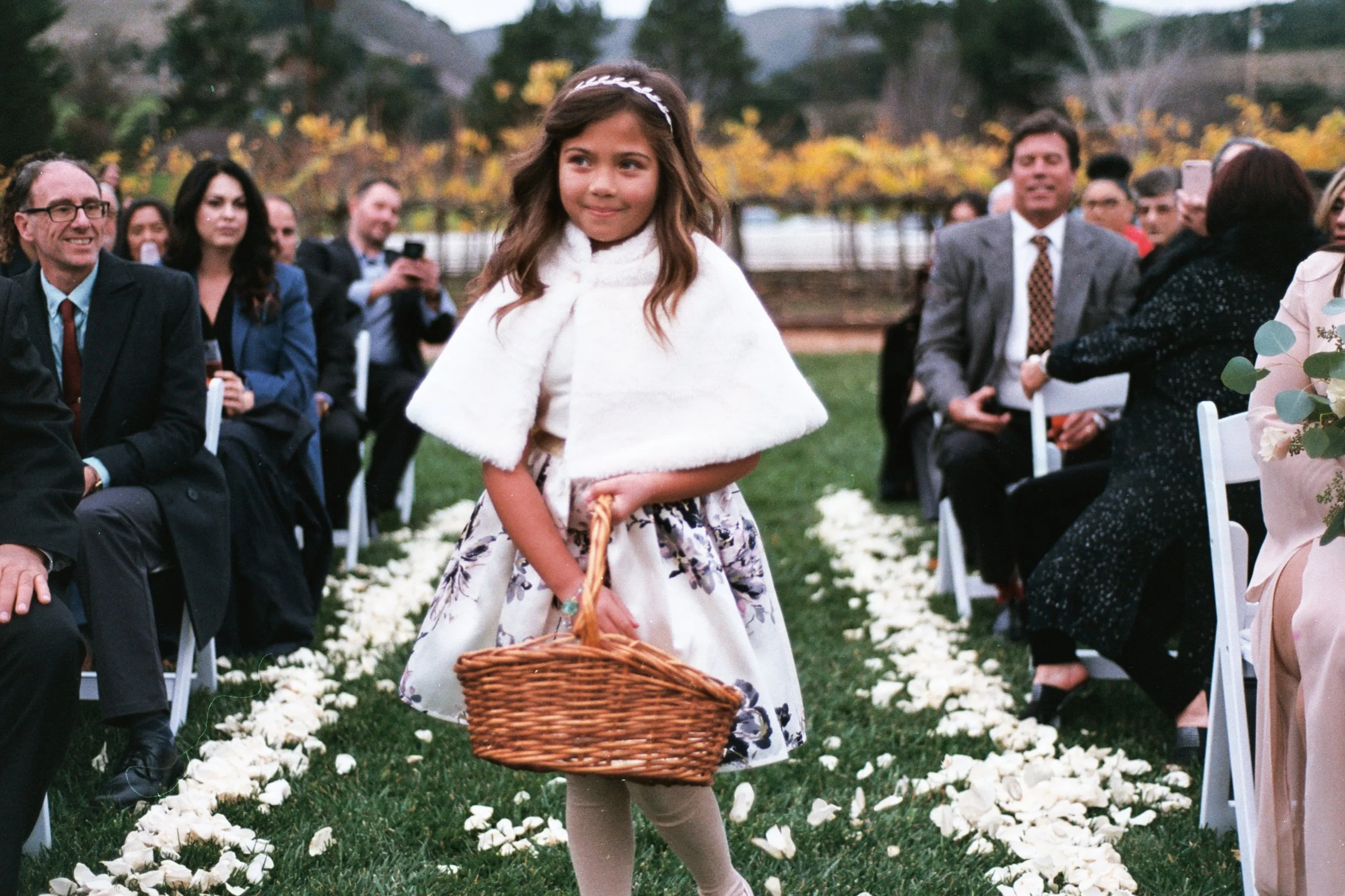 A girl dressed in formal attire with a white fur cape and floral dress, holding a basket, walking down an aisle decorated with white flowers at an outdoor event, surrounded by seated guests in suits and dresses.