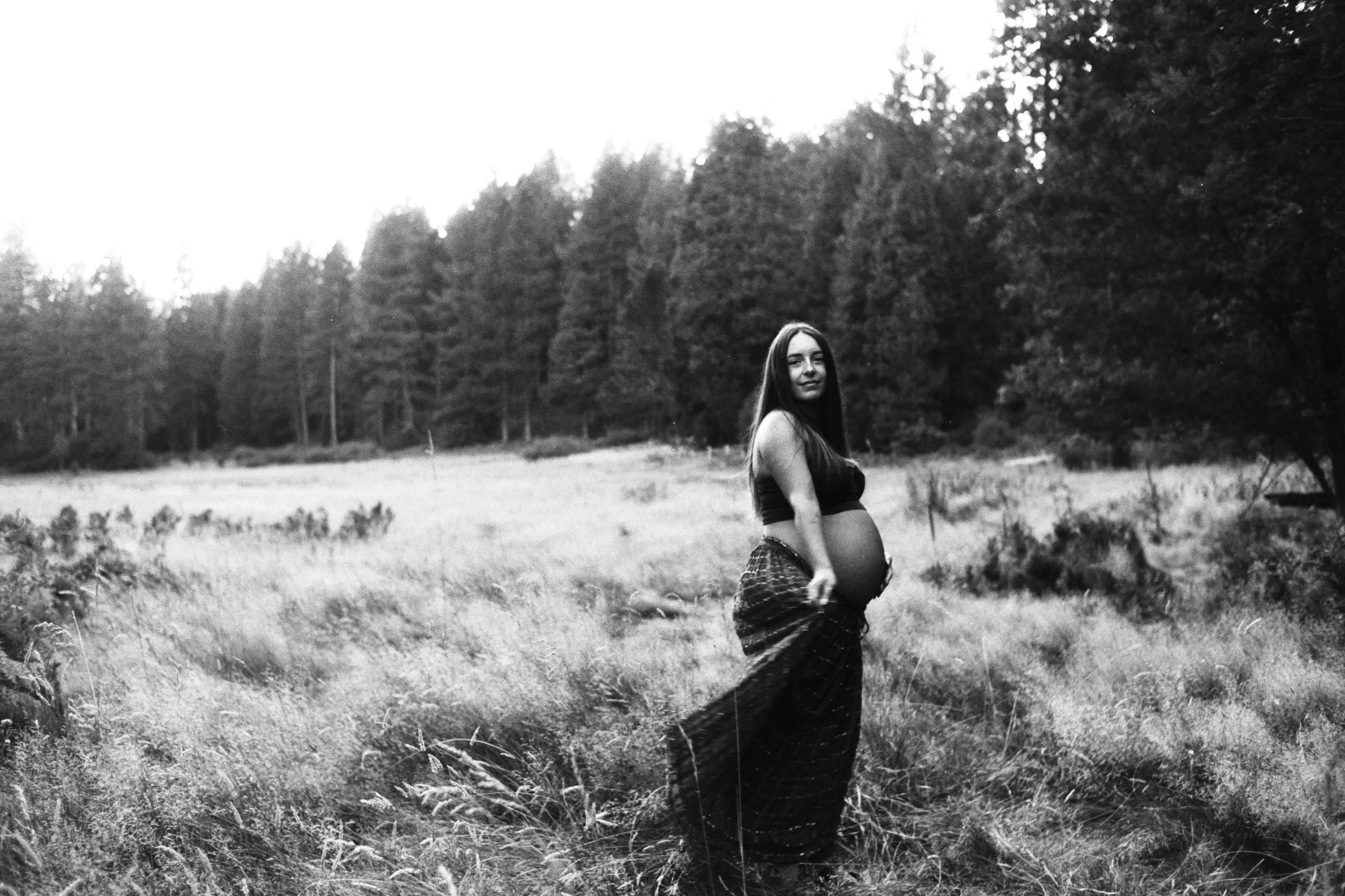 A pregnant woman standing outdoors in a field with trees in the background, in black and white.