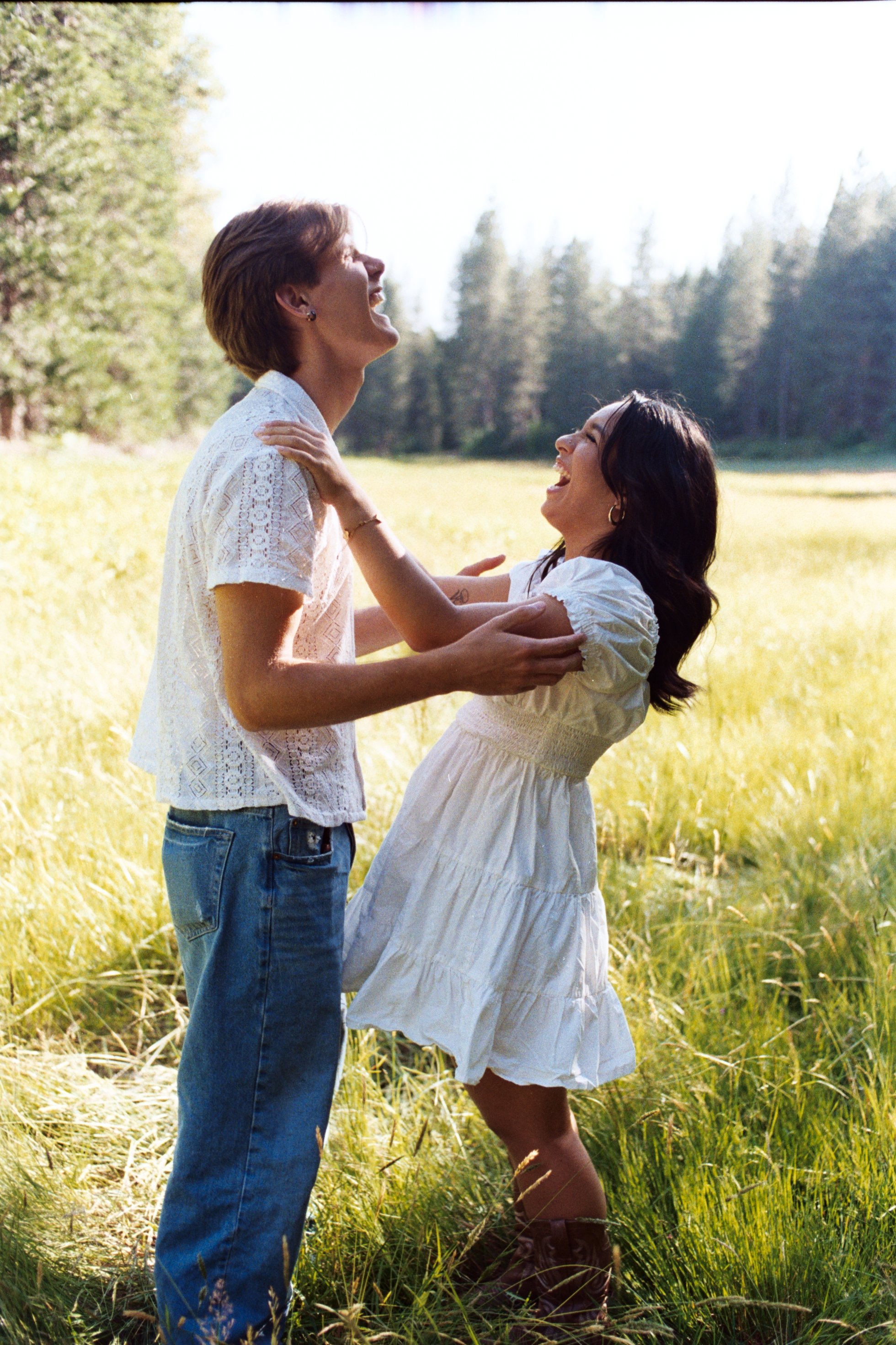 A man and a woman are enjoying a joyful moment outdoors in a grassy field with trees in the background. The man is wearing a white lace shirt and blue jeans, and the woman is wearing a white dress and brown boots. They are smiling and holding onto ea