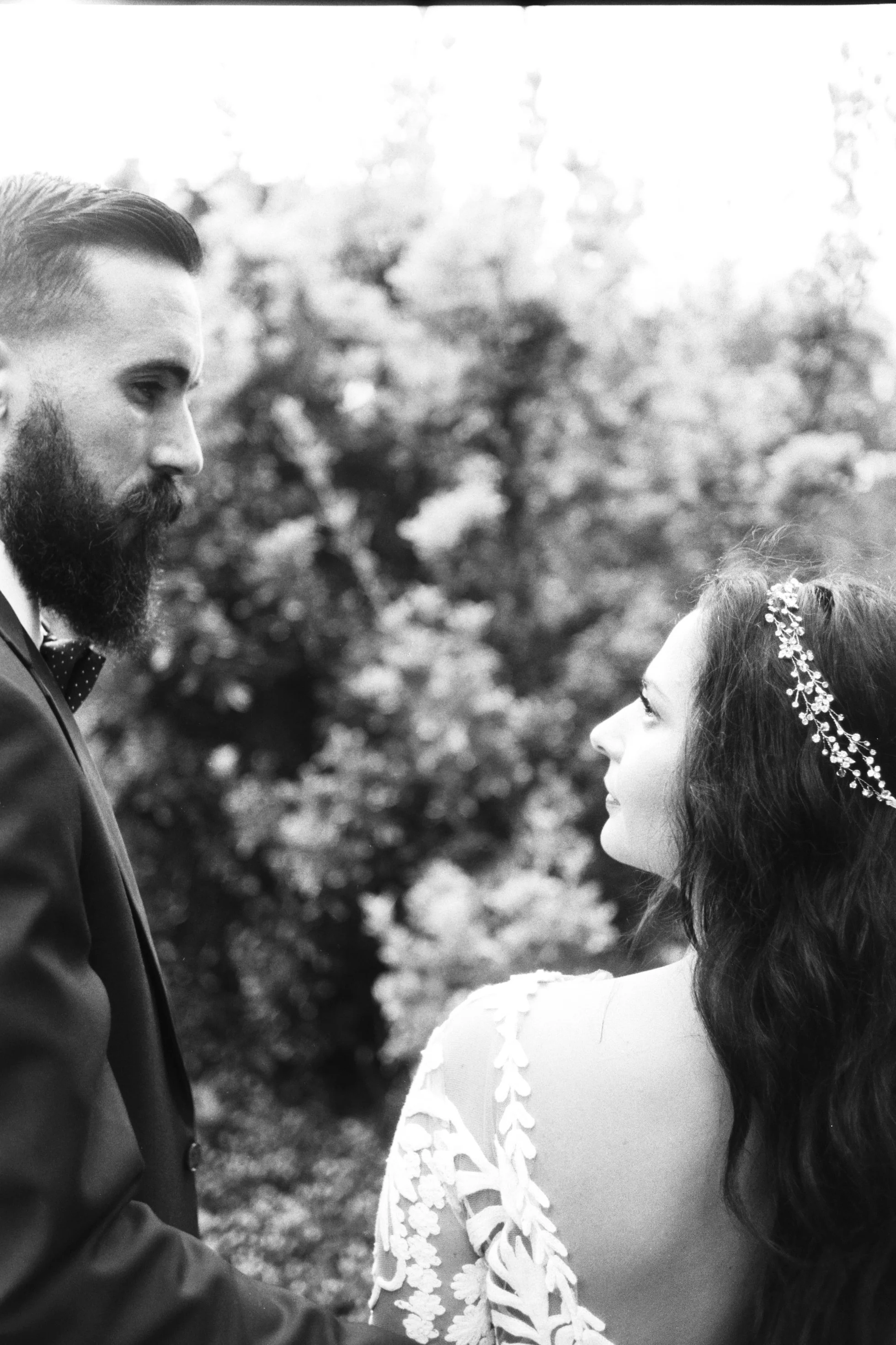 A black and white photo of a man and woman, facing each other outdoors. The man has a beard and is wearing a suit, and the woman has long hair with a floral headband. They are looking into each other's eyes.