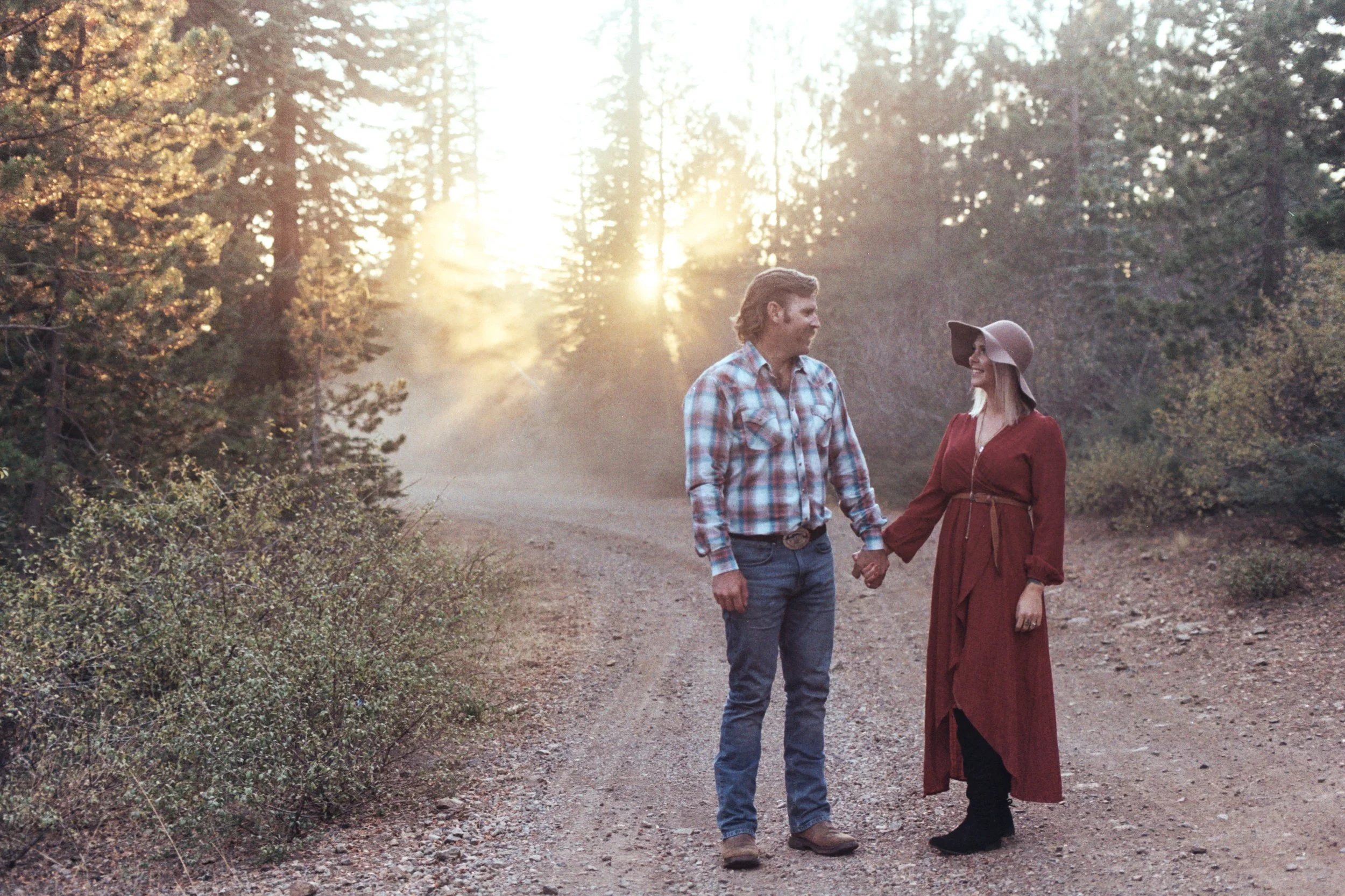 A man and woman holding hands and walking on a dirt path in a forest during sunset, with trees and sunlight in the background.