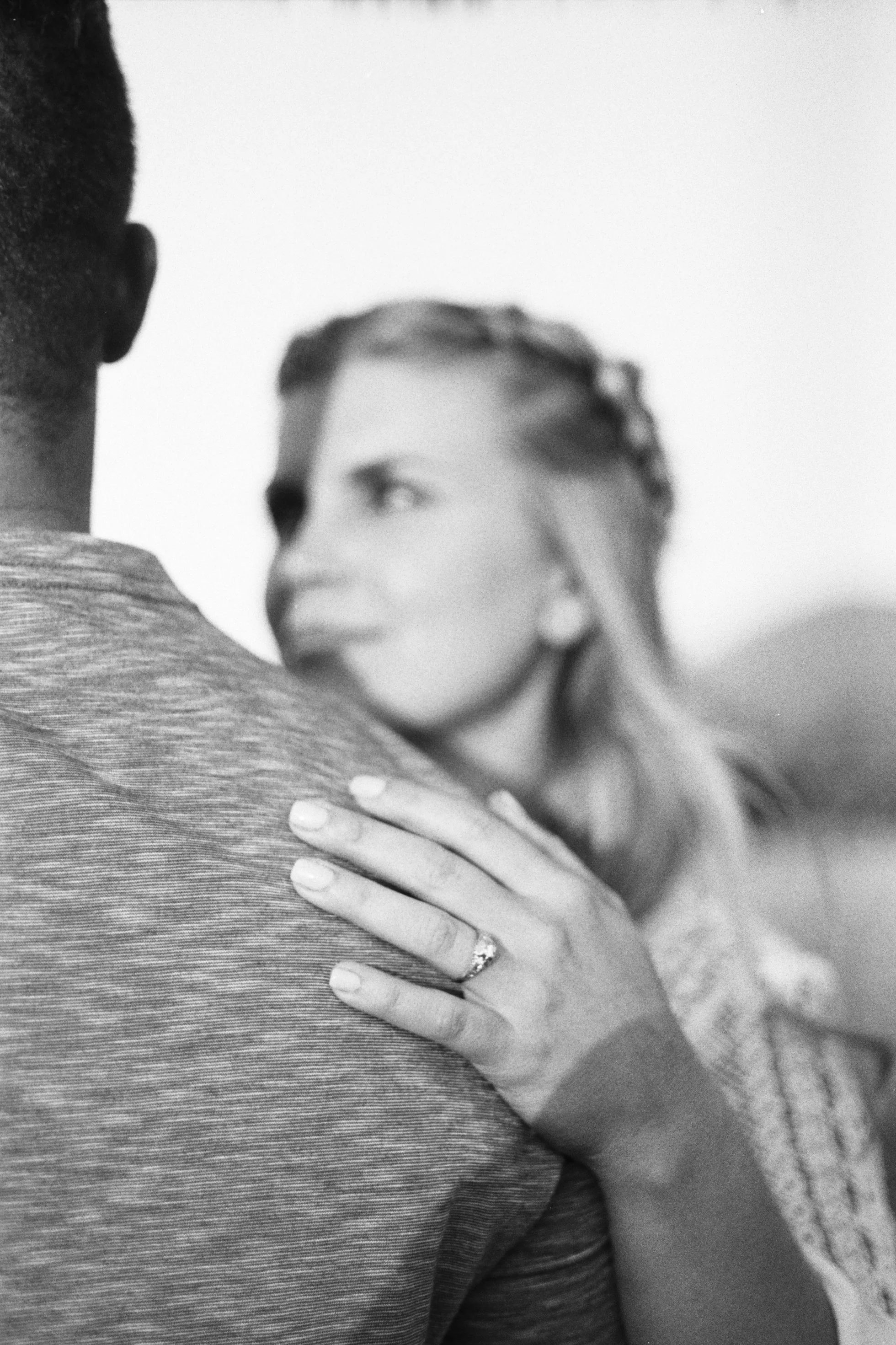A woman with light skin and curly hair is embracing a man with dark skin, whose back is to the camera. The woman has her hand on the man's shoulder, and she is wearing a ring on her finger, suggesting a romantic relationship.