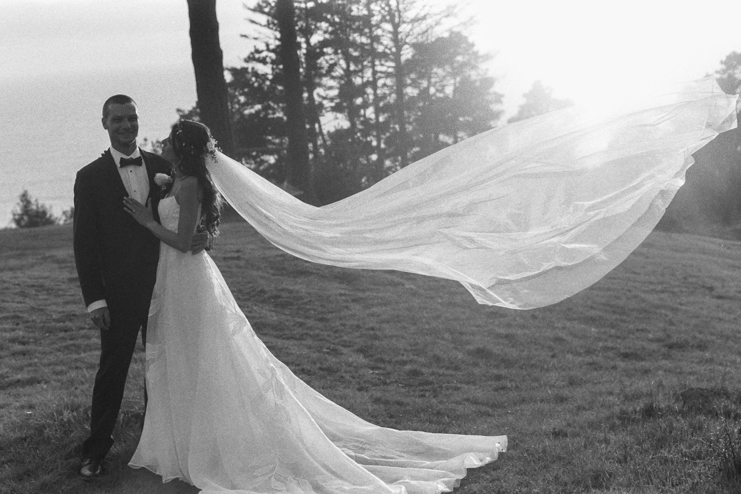 A black and white photo of a bride and groom dancing outdoors, with the bride's veil flowing behind her and trees in the background.