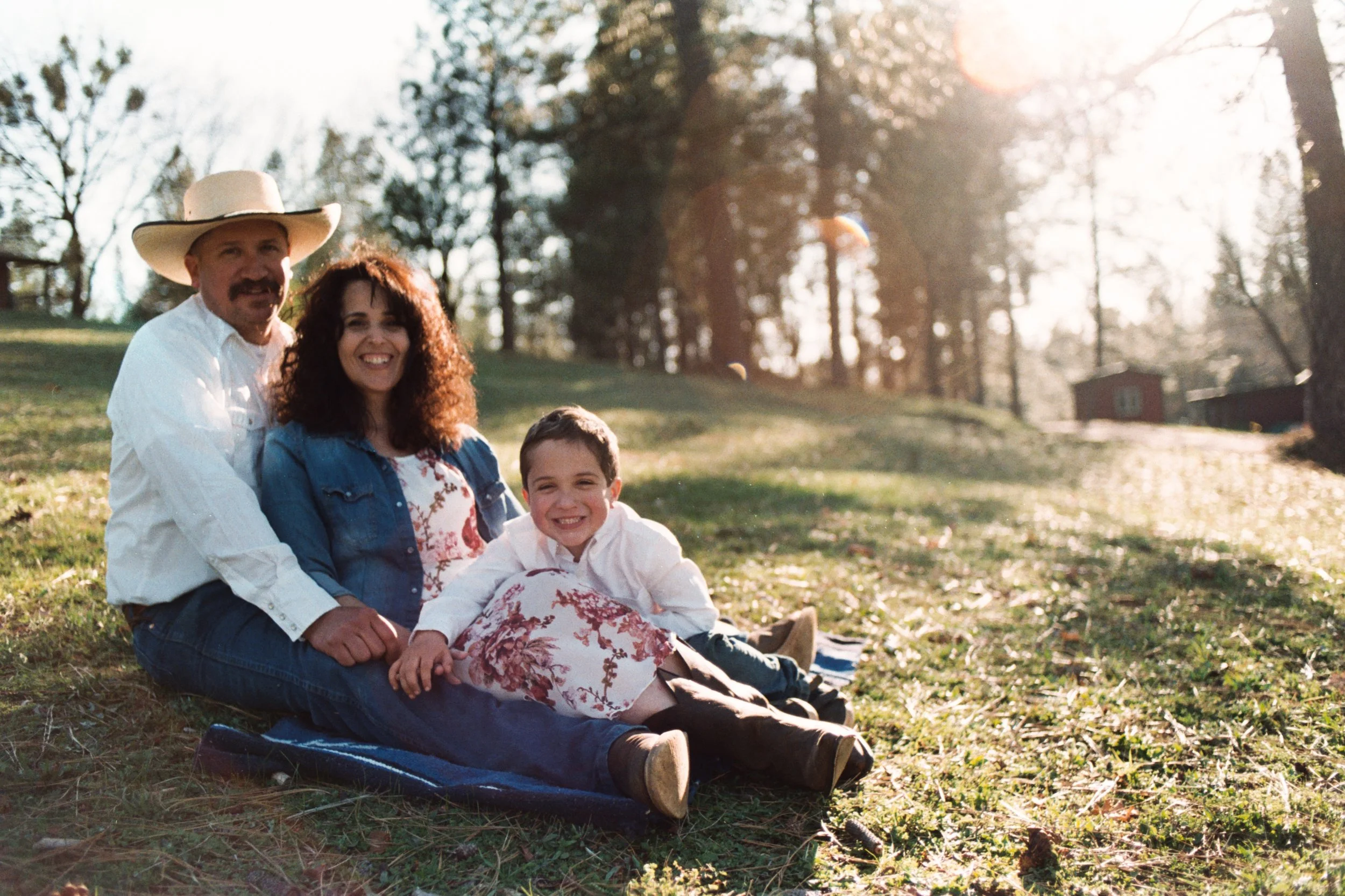A family of four sitting on a blanket outdoors in sunlight with trees and a house in the background, smiling and enjoying the day.