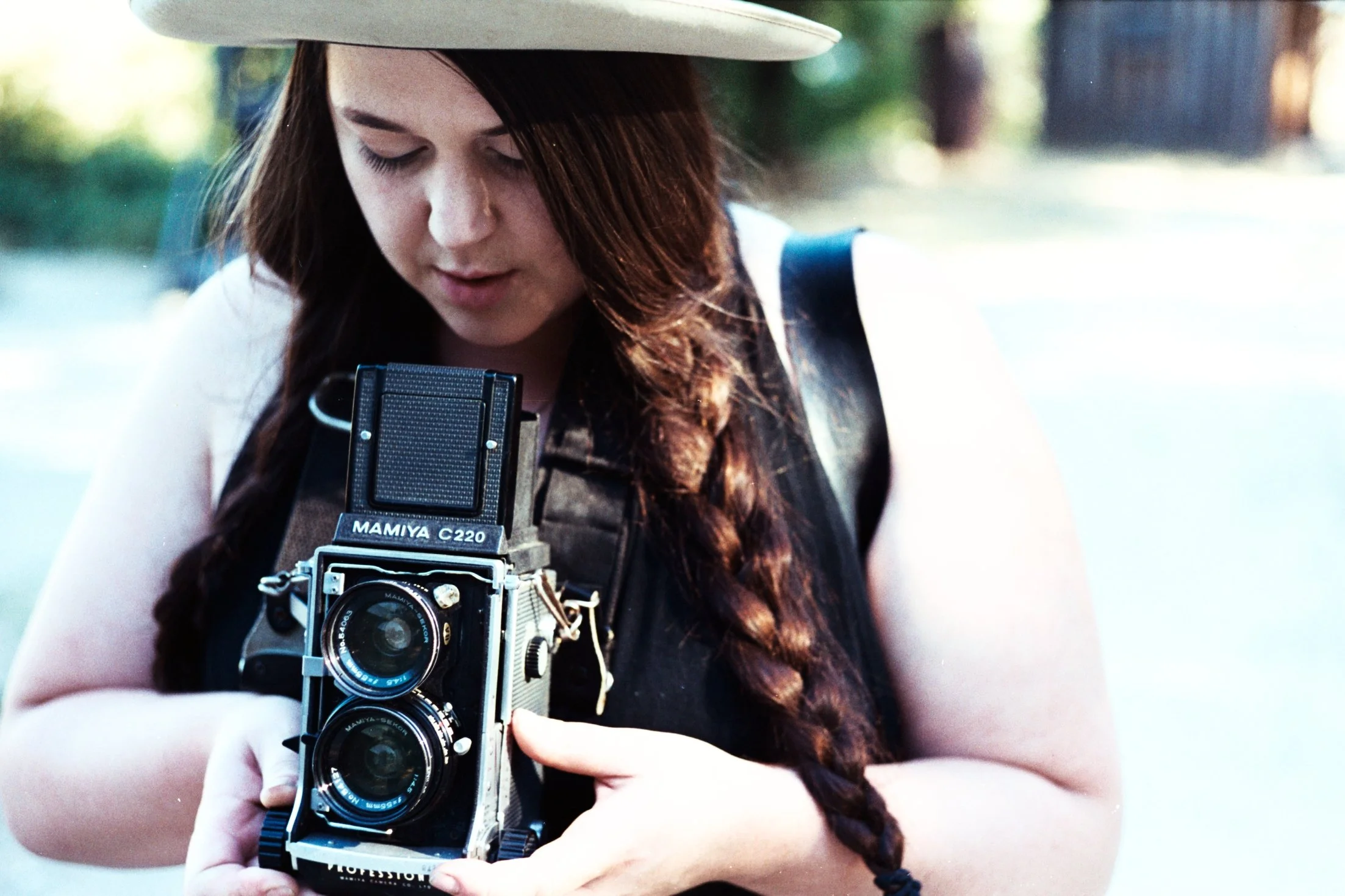 A woman with long hair wearing a wide-brimmed hat and a vest, holding a vintage camera outdoors.
