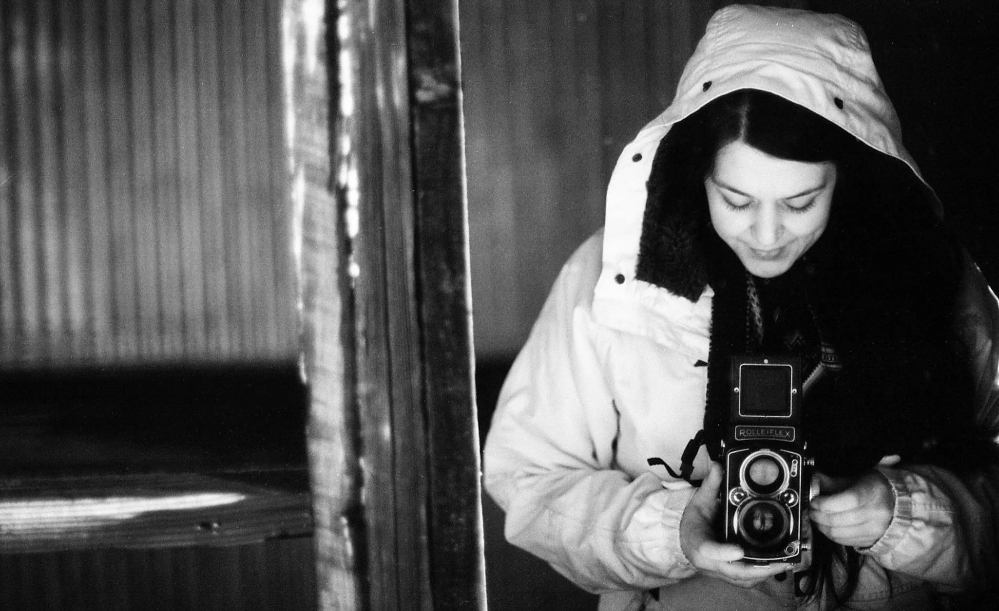 A woman wearing a hooded jacket taking a self-portrait with a camera, with her head bowed and eyes closed, in black and white.