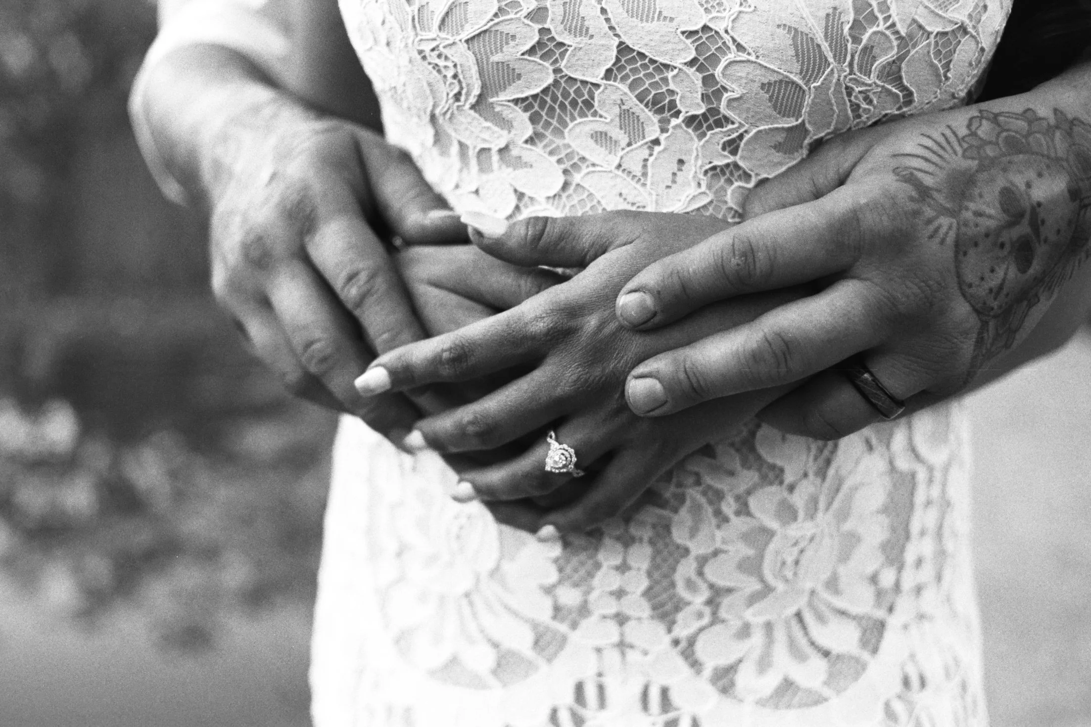 A close-up black and white photo of two hands clasped together, one hand wearing a wedding ring, resting on the lace fabric of a person's dress.