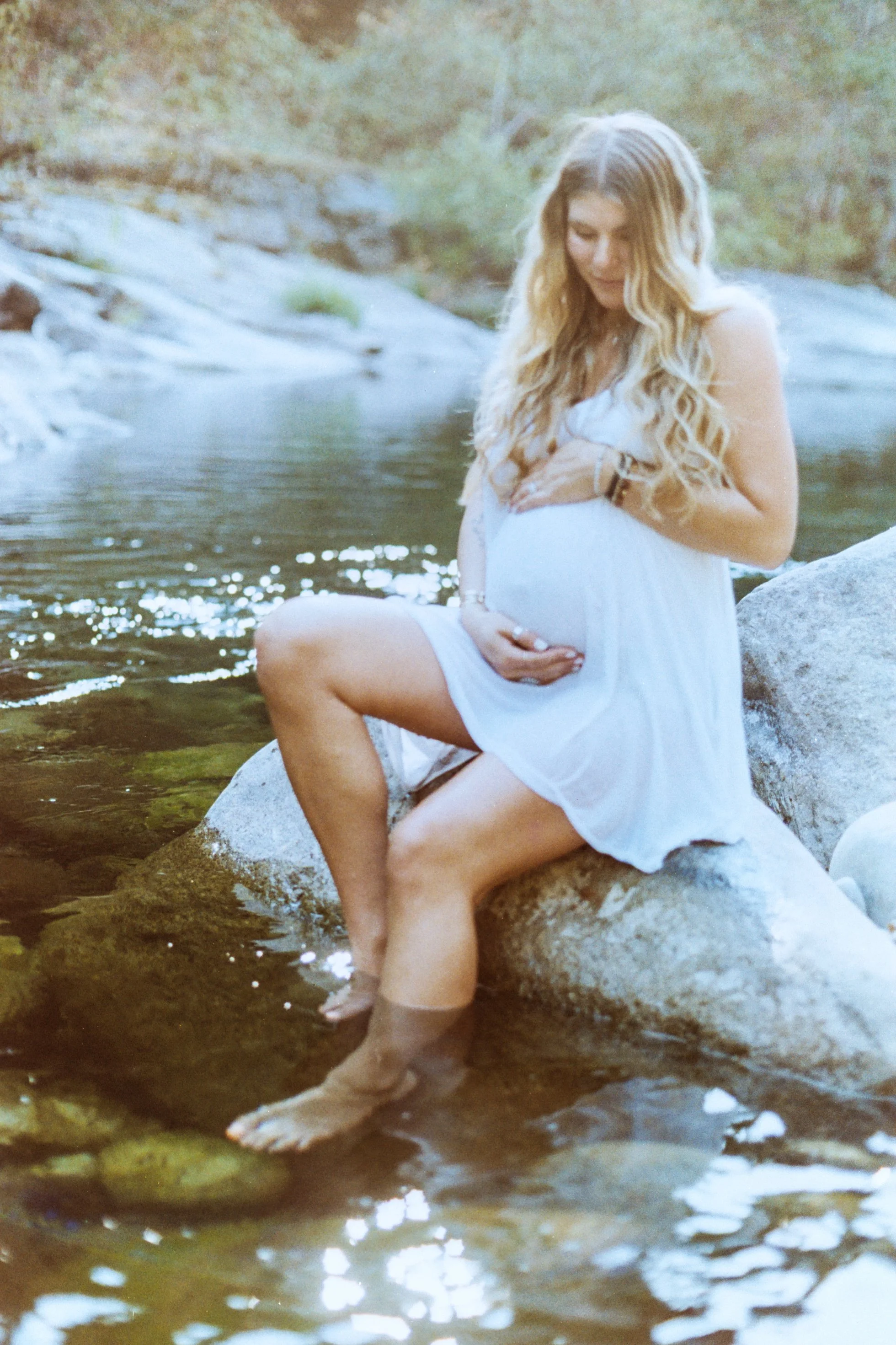 A woman with long, wavy blonde hair sitting on a rock by a river, with her hand on her pregnant belly, looking down with a gentle smile.