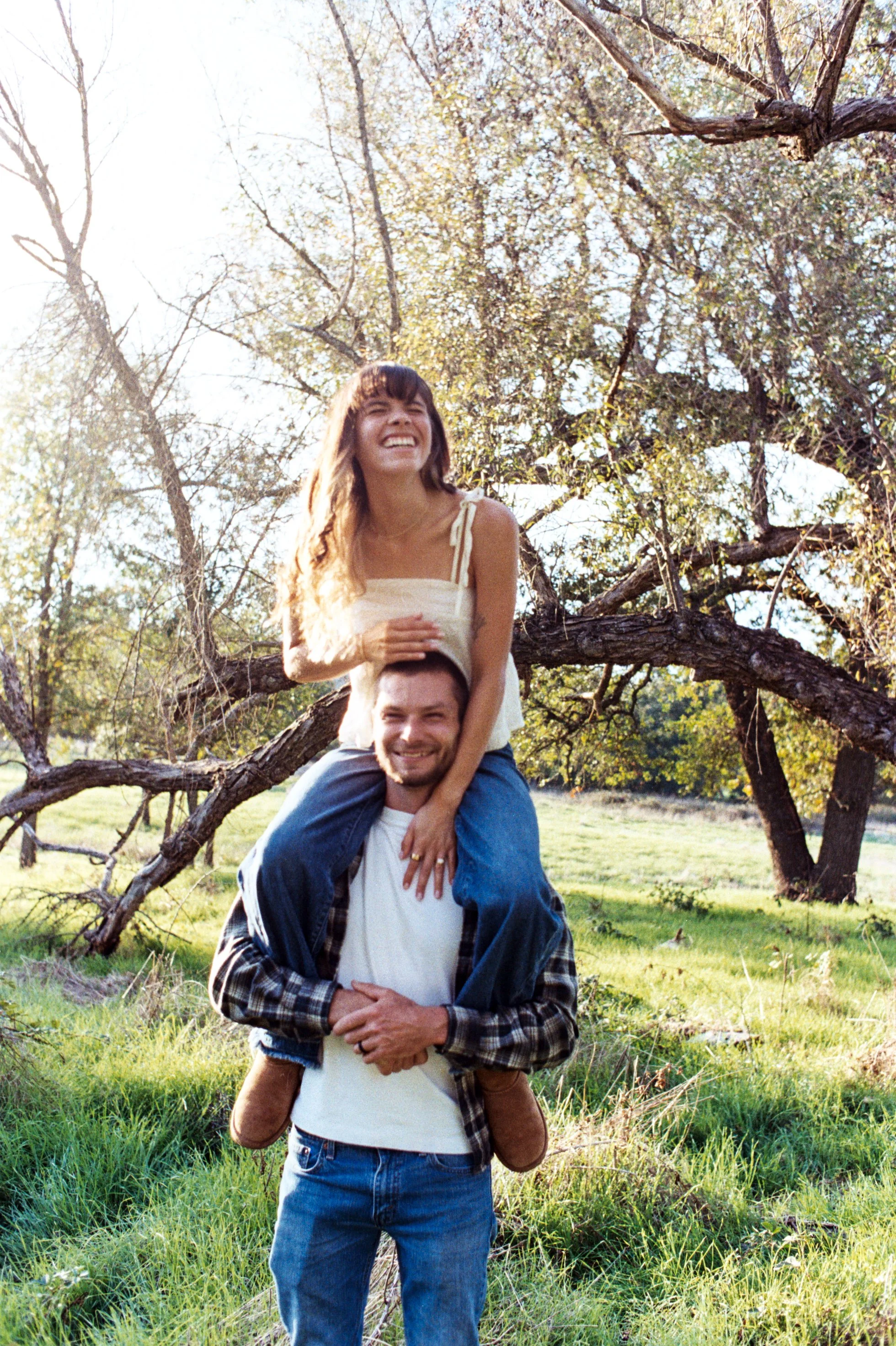 A young woman with long hair and a white dress is sitting on a man's shoulders, both smiling, outside near a tree with sprawling branches in a grassy field, during daytime.