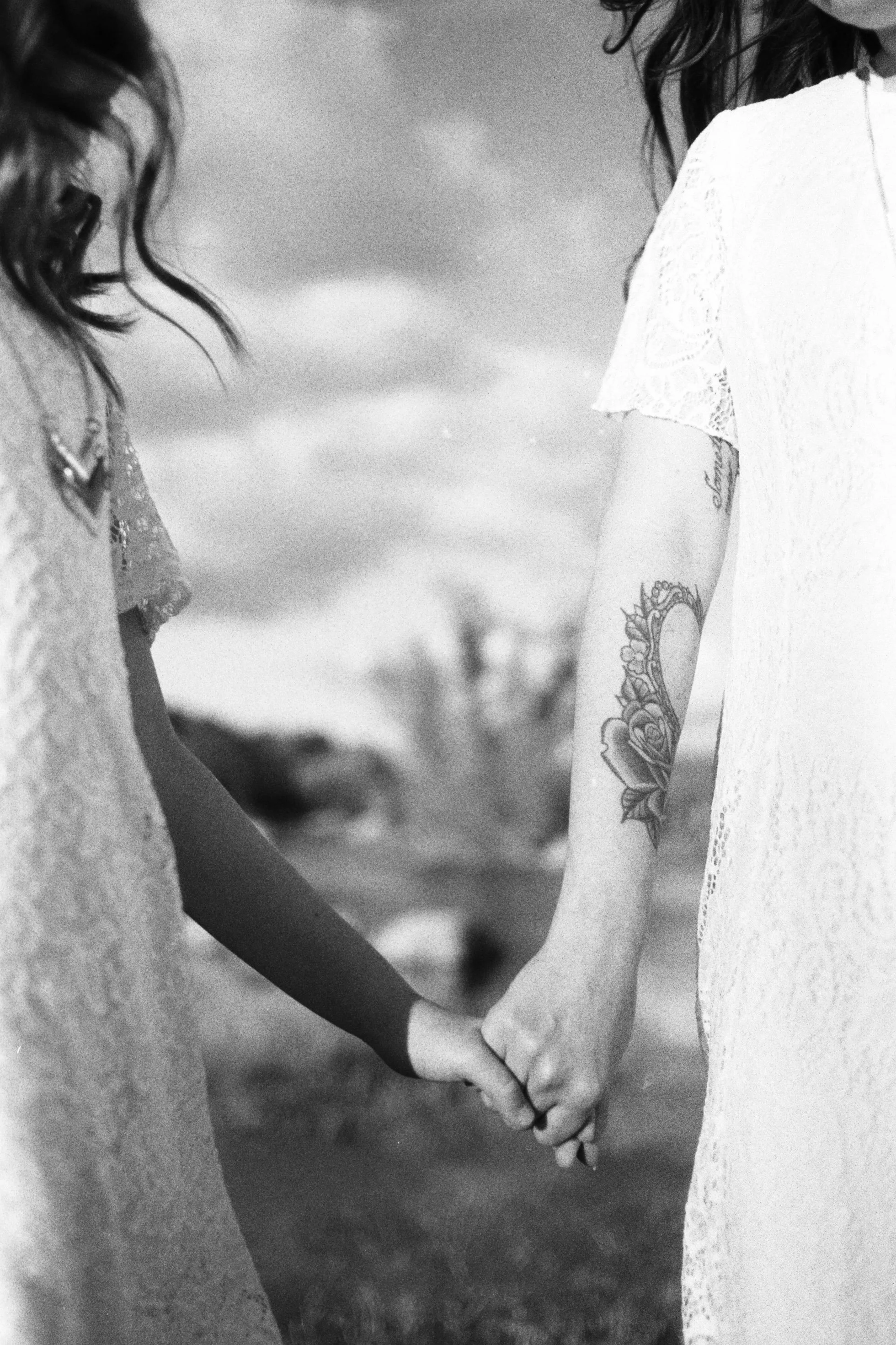 Two people holding hands, close-up of their arms and intertwined fingers, one person has a tattoo of a flower on the forearm, both are wearing light-colored clothing.
