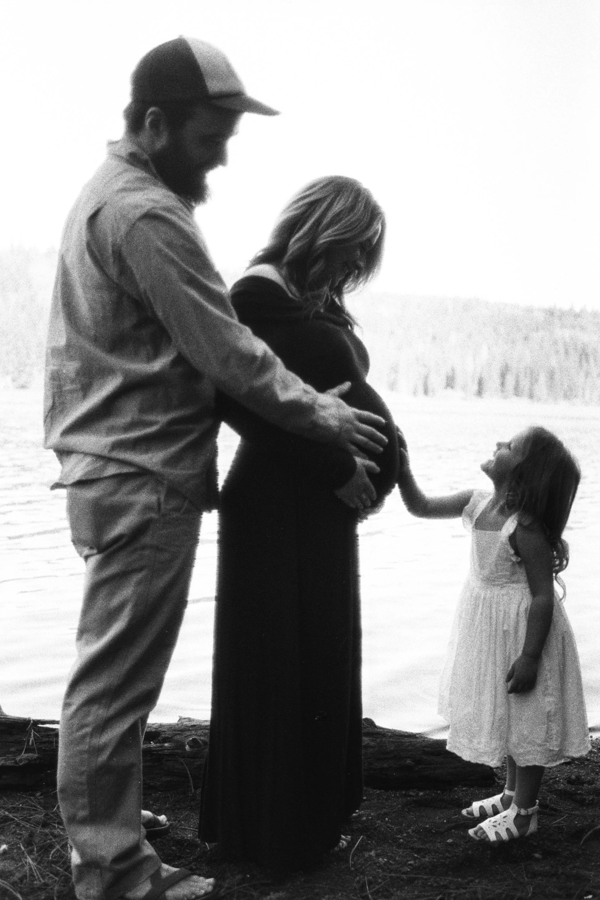 A family of four standing outdoors near a body of water. The father and mother are holding their young children, a boy and a girl, as they look at each other and smile.