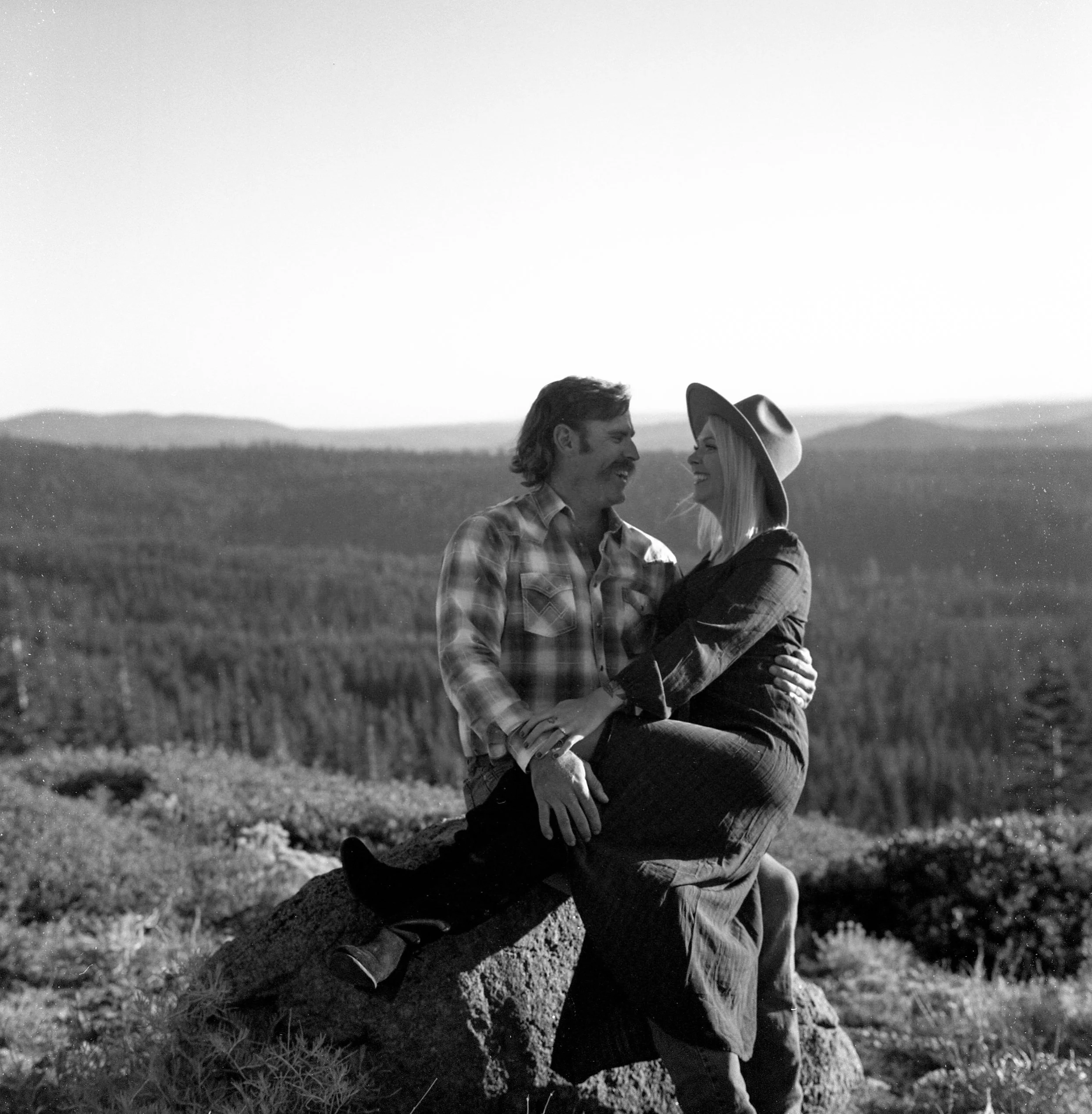 A black and white photo of a smiling man and woman in an outdoor rural setting, possibly a farm, with the man sitting on a stone and the woman standing next to him. The woman is wearing a wide-brimmed hat and they are looking at each other.