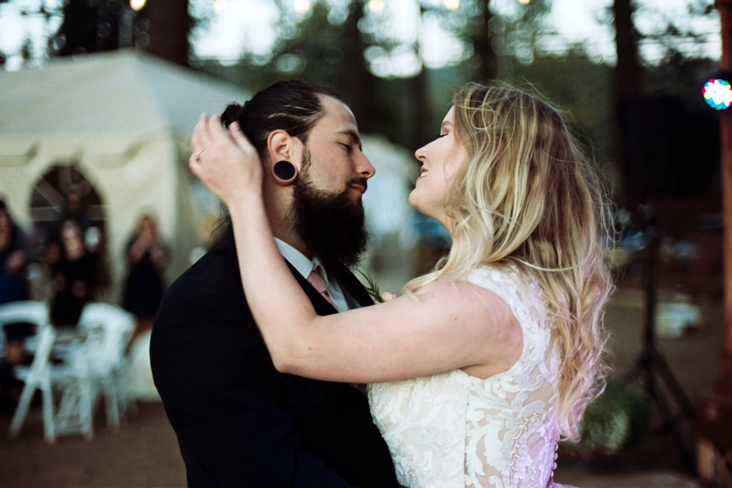 A couple dancing closely at a wedding reception, with the woman wearing a white lace dress and the man in a black suit. They are outdoors with trees and blurred guests in the background.