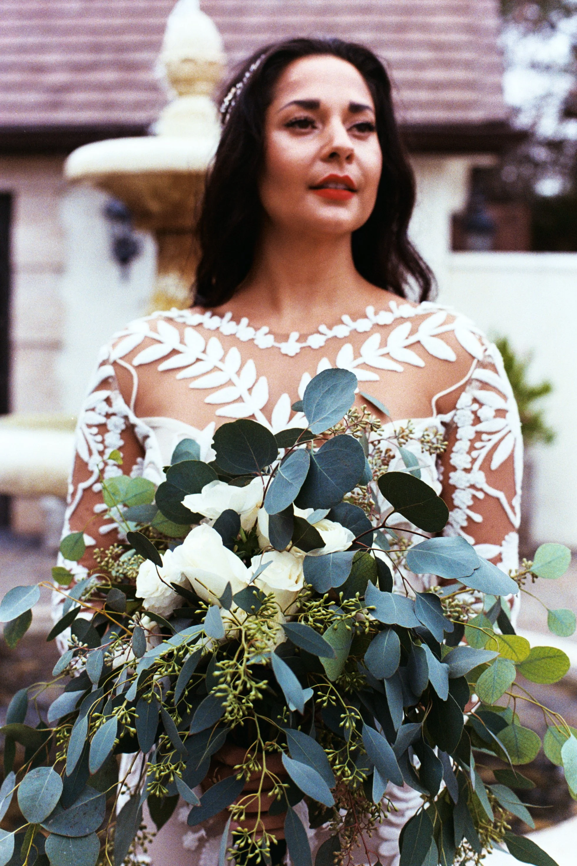 A bride holding a bouquet of white flowers and greenery, with an outdoor fountain and house in the background.