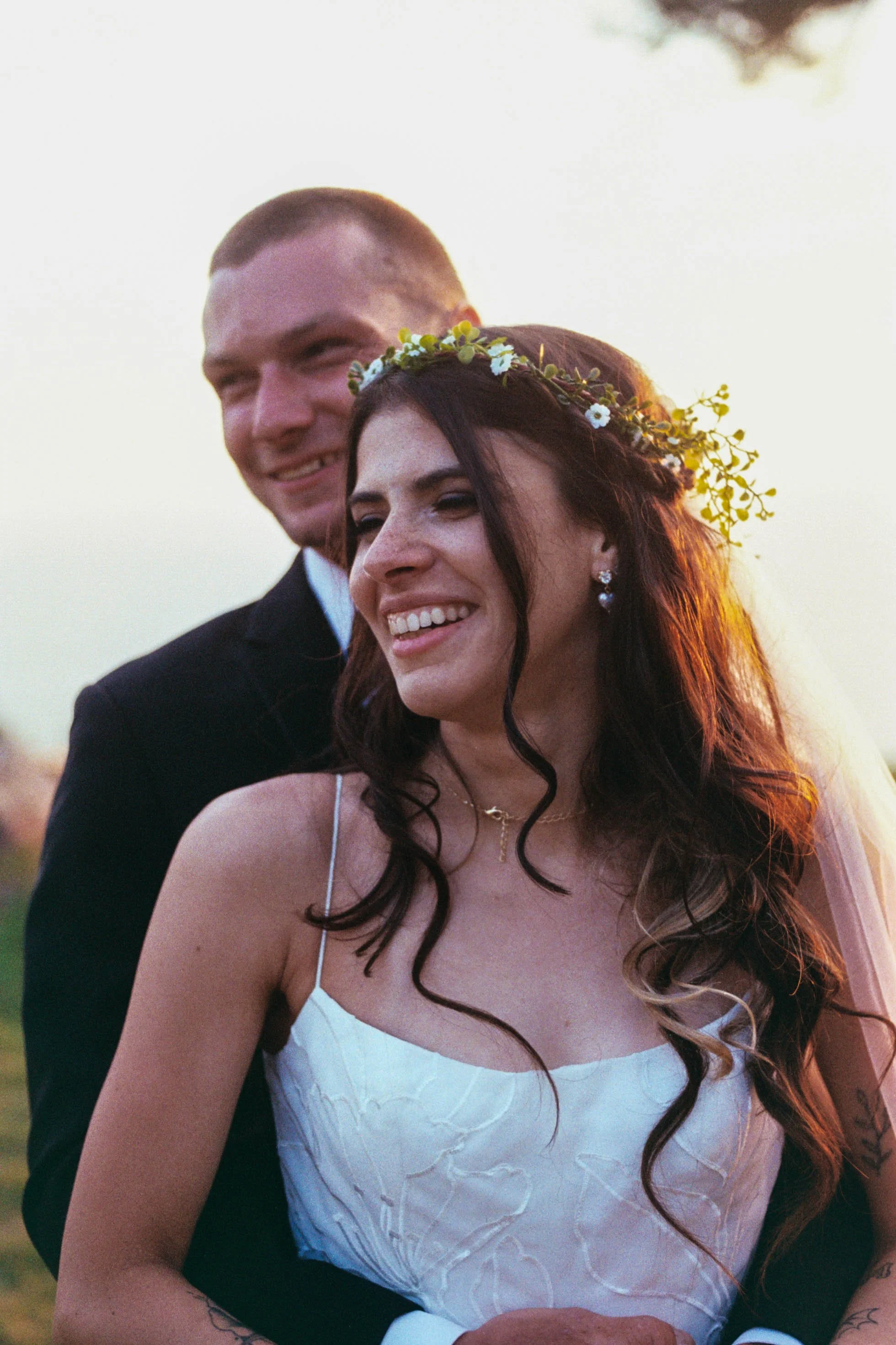 A smiling couple at sunset, with the woman wearing a white dress and a flower crown, and the man in a black suit with a white shirt.
