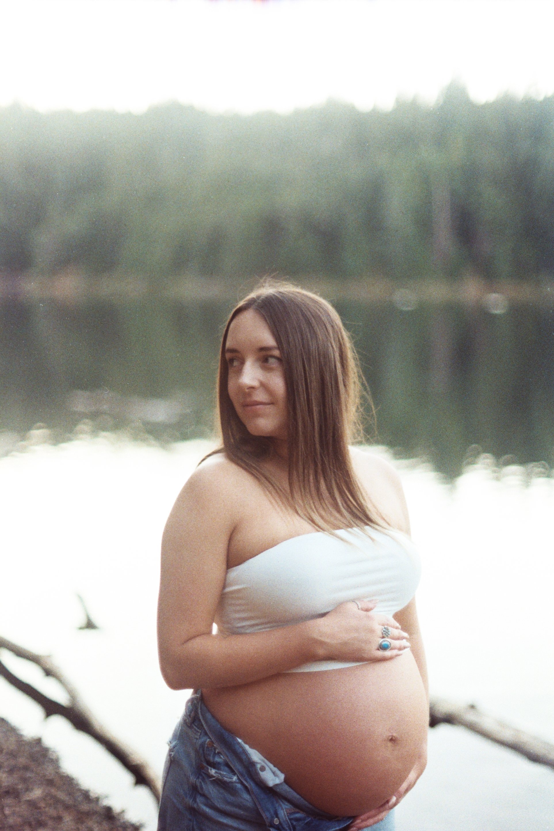 A pregnant woman with long brown hair, wearing a white tube top and denim shorts, standing outdoors near a body of water with trees in the background.
