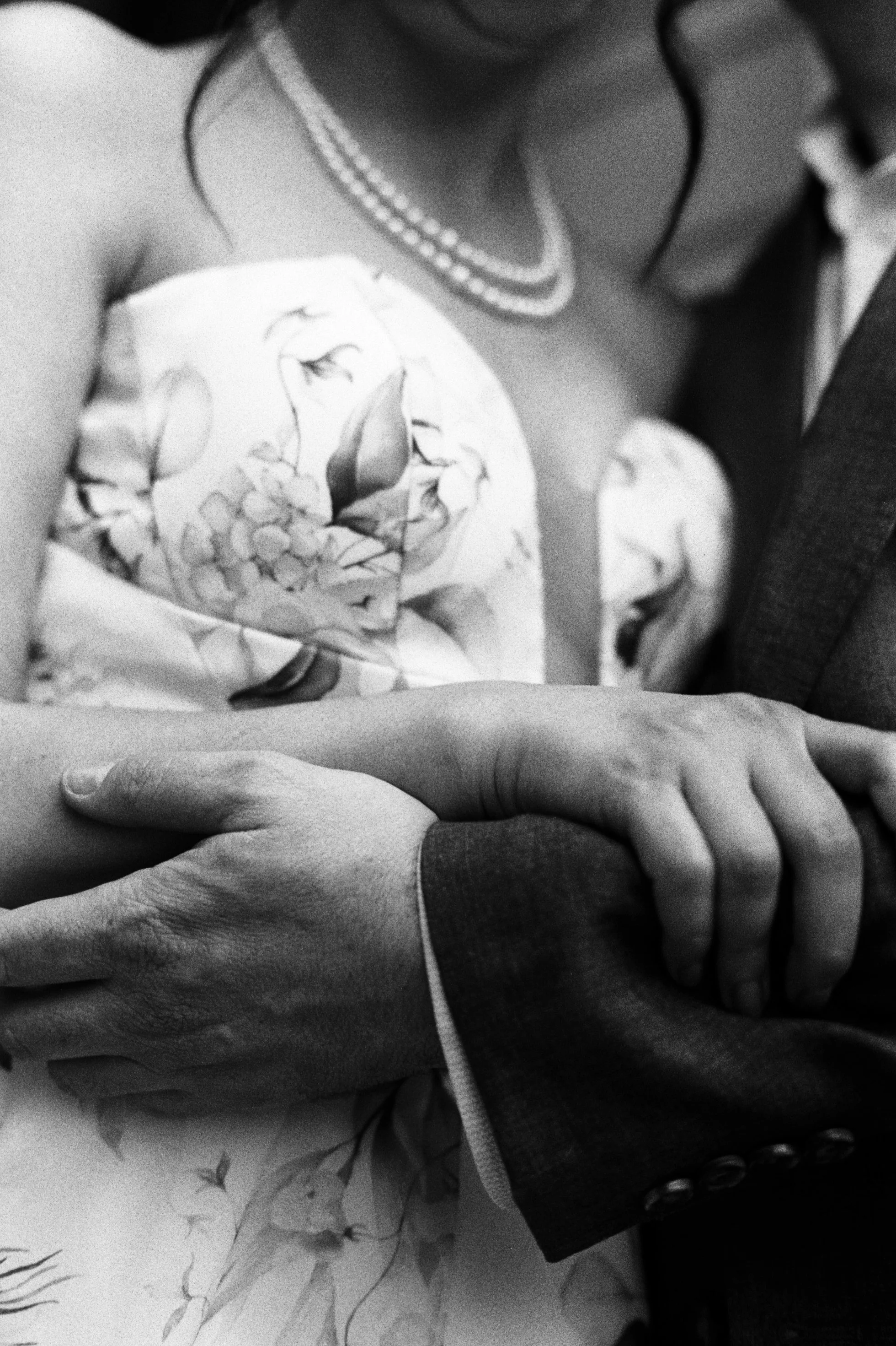 Close-up of a couple holding hands, with one person wearing a layered pearl necklace and a floral dress, and the other wearing a dark suit sleeve.