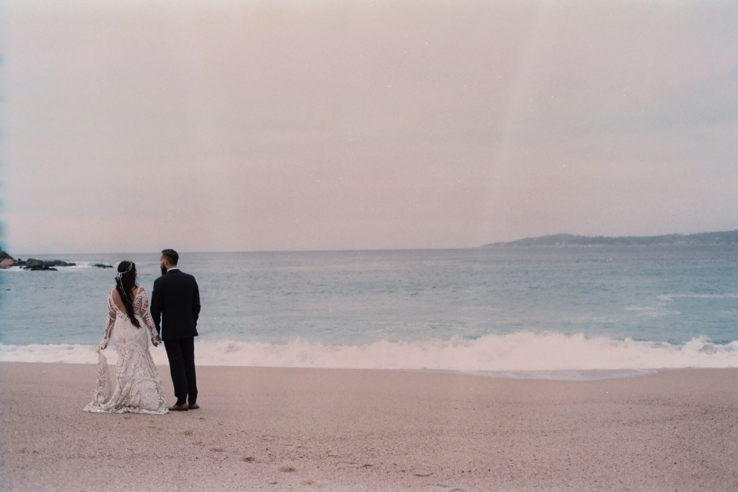 A bride and groom standing on the beach near the water with a scenic view of the ocean and distant land, possibly during their wedding or vow renewal.