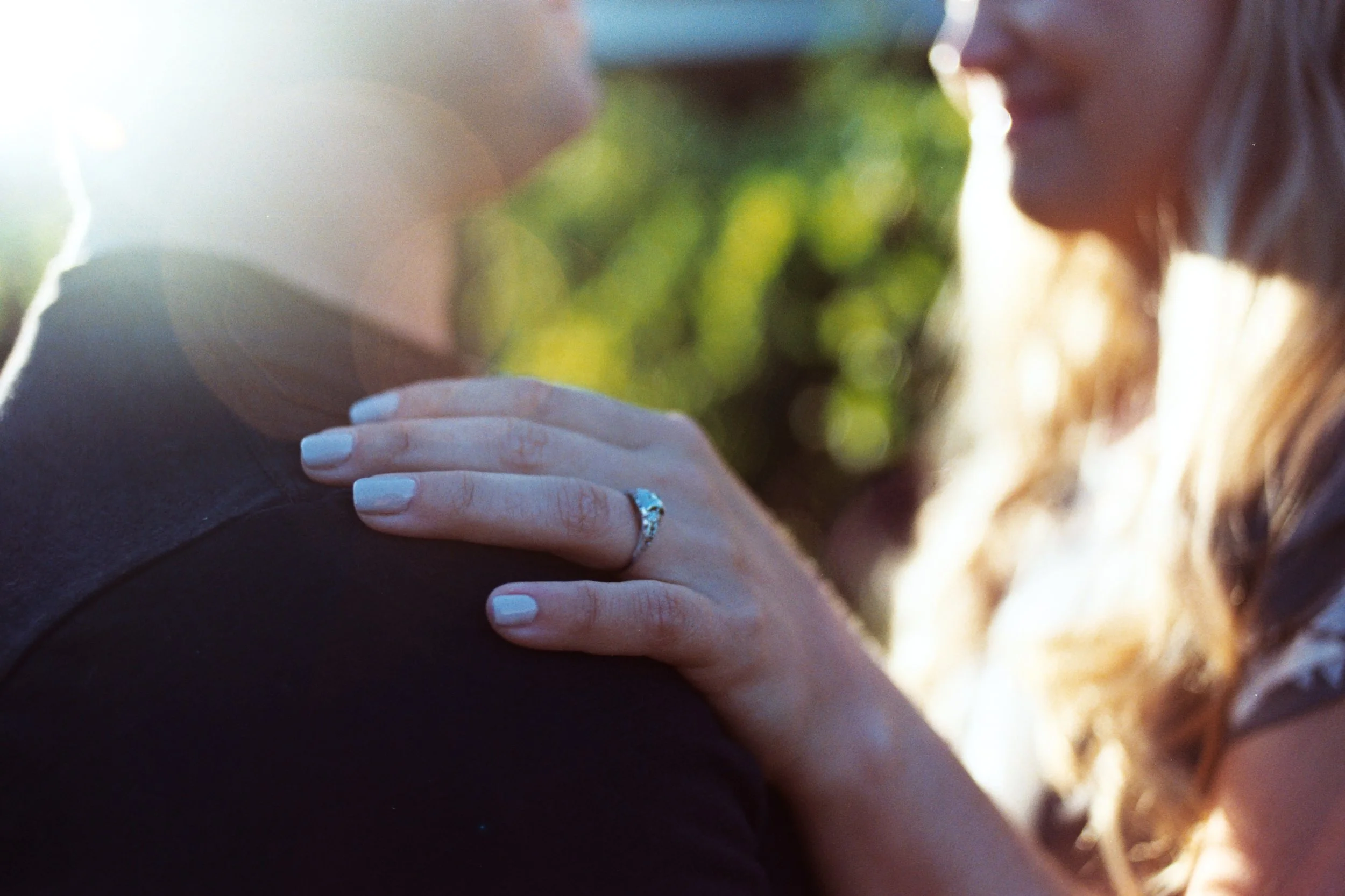 Close-up of a woman with light skin, blonde hair, and white nails touching someone's shoulder, with a wedding ring visible on her finger, during sunset outdoors.