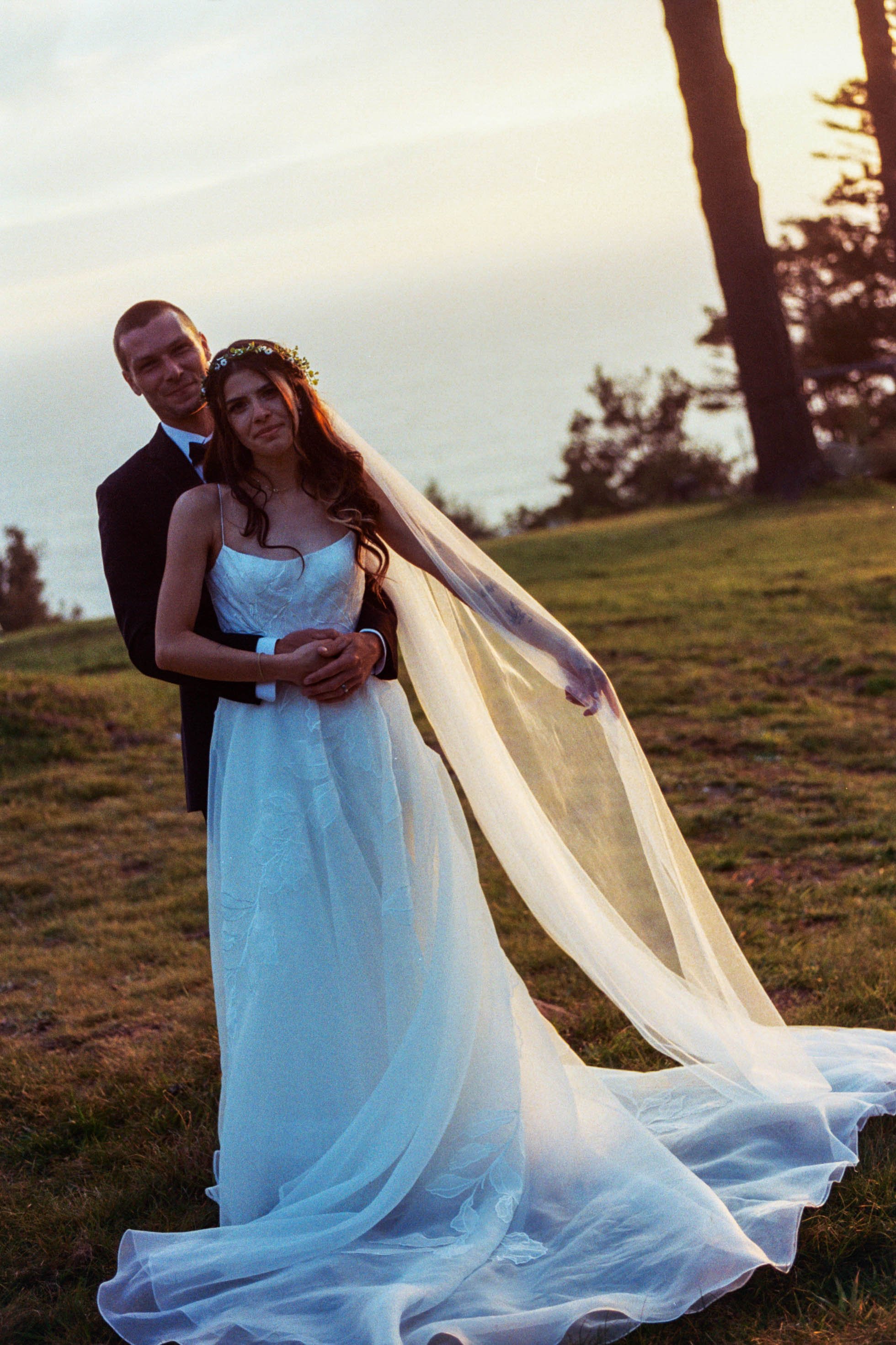 A bride and groom standing together outdoors during sunset, with the bride wearing a white wedding dress and veil, and the groom in a black tuxedo. The bride has a flower crown and the veil flows behind her, with trees and a grassy area in the backgr