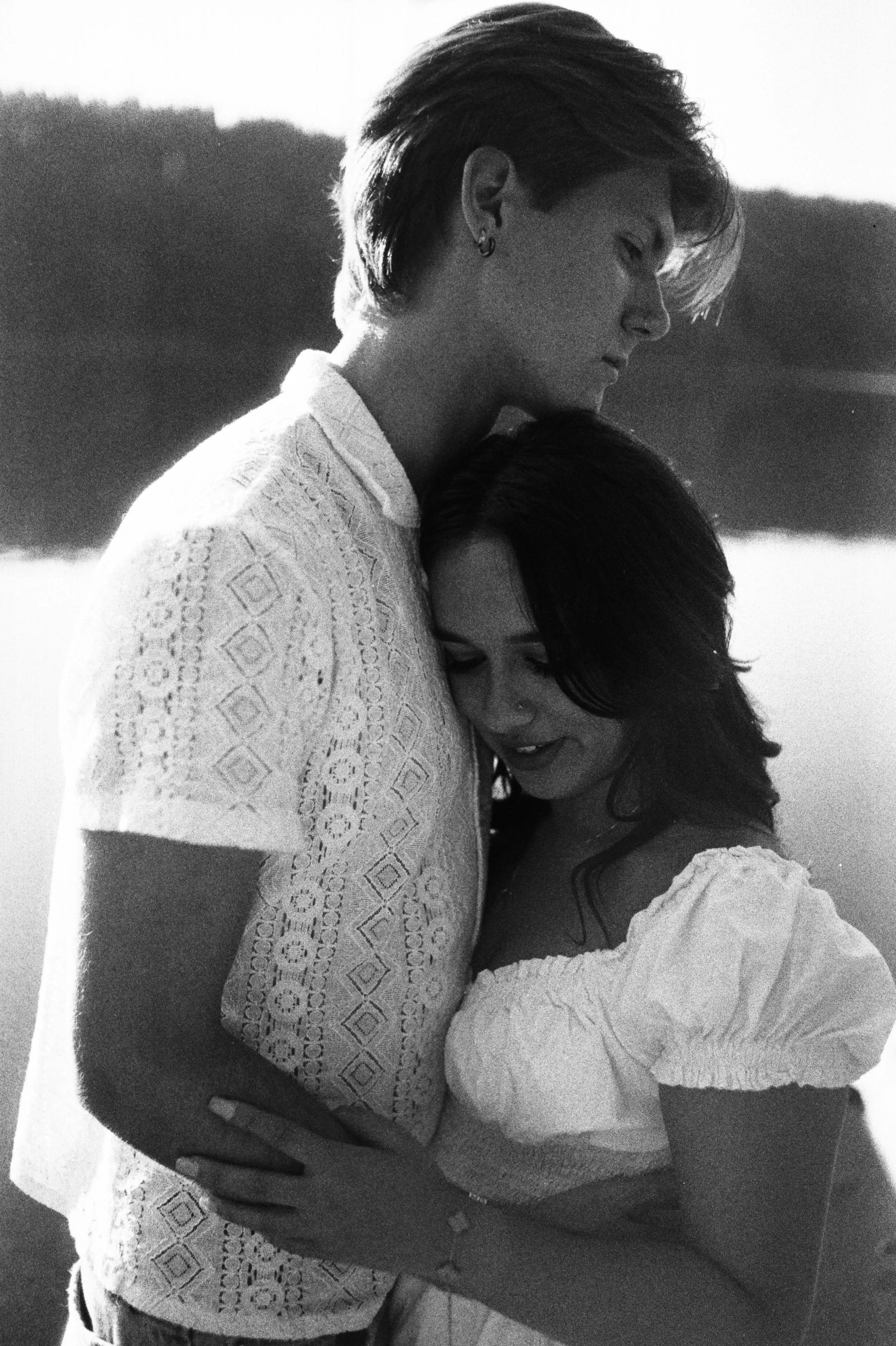 A black and white photo of a woman with short hair and an earring, embracing a person with their face in her chest, both wearing light-colored dresses with textured patterns. The woman looks down with a gentle expression, and the background is blurre