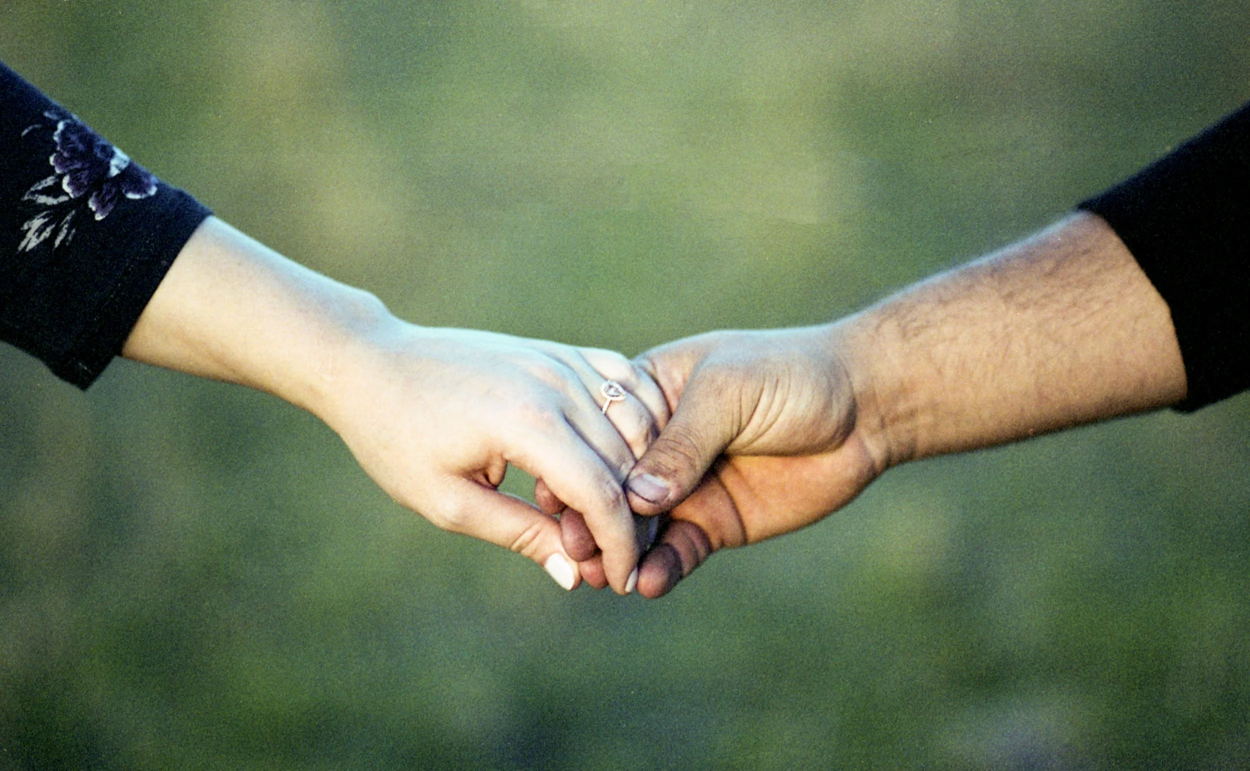 Close-up of a Caucasian woman and a person of color holding hands with interlocked fingers, against a blurred outdoor background.