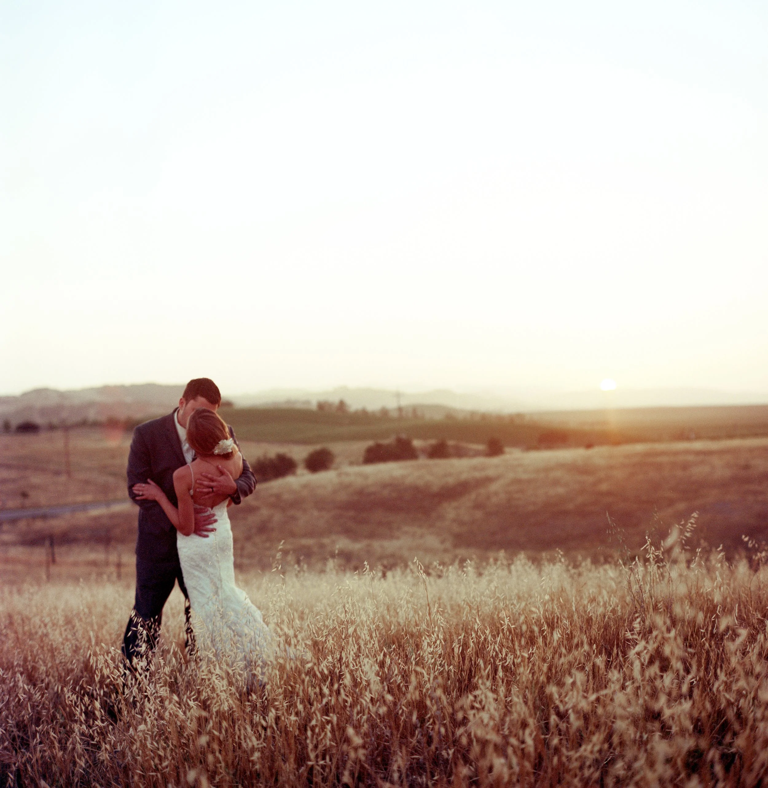 A couple, dressed in wedding attire, embracing in a field of tall grass during sunset with rolling hills in the background.