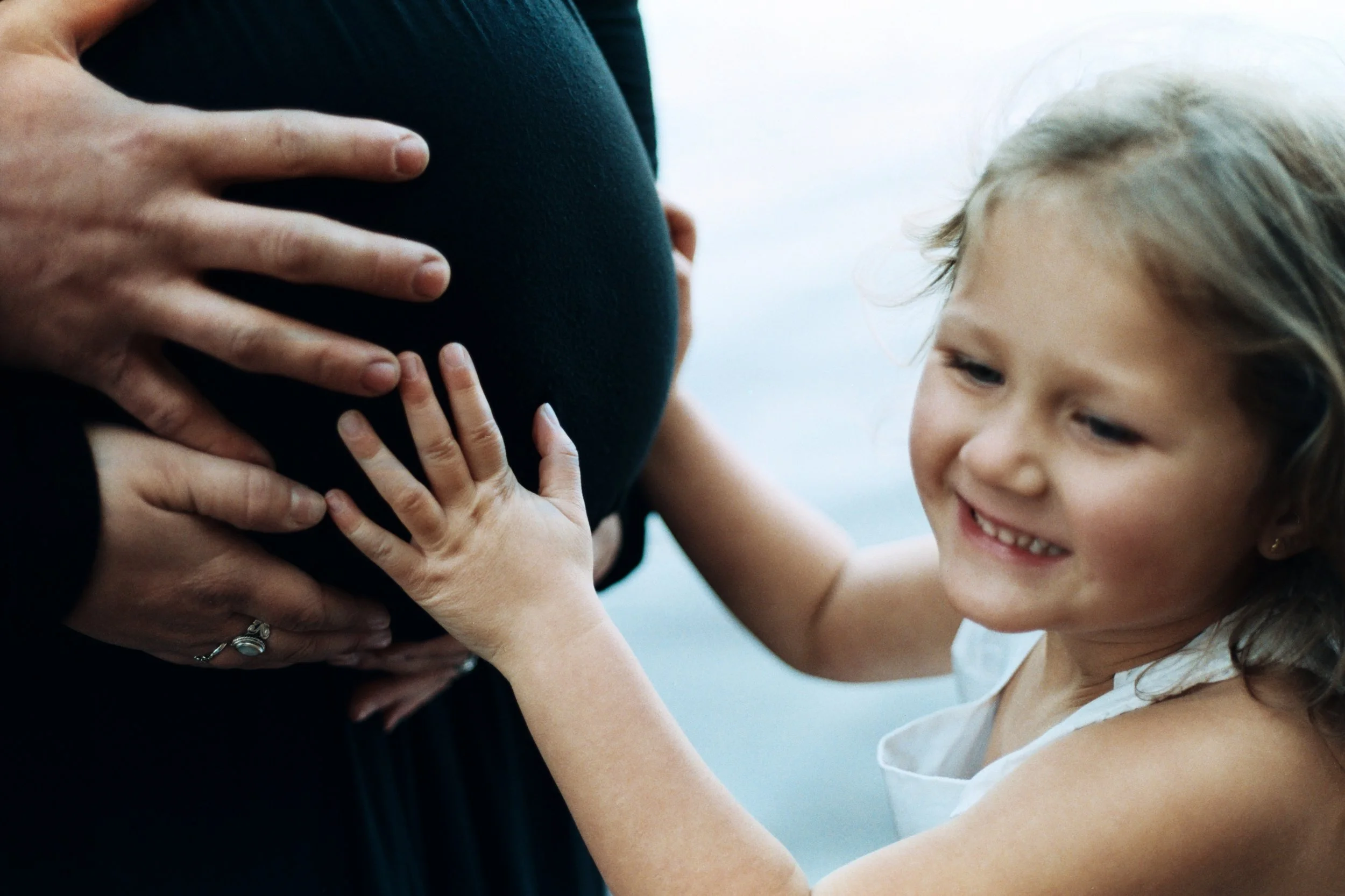 A young girl with blonde hair smiling and touching a pregnant woman's belly with her hands. The woman is wearing a dark shirt and her hand is resting on her belly.