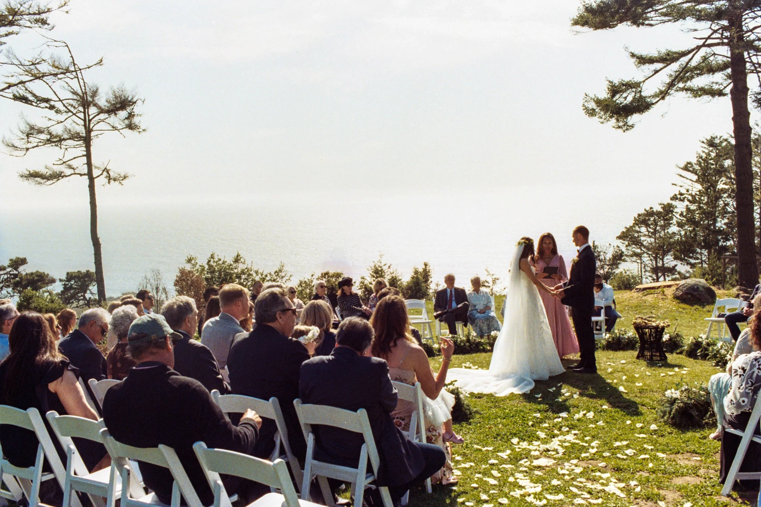A wedding ceremony taking place outdoors on a grassy area, with the bride and groom standing together, surrounded by guests seated in white chairs. Tall trees and a scenic view of water are visible in the background.