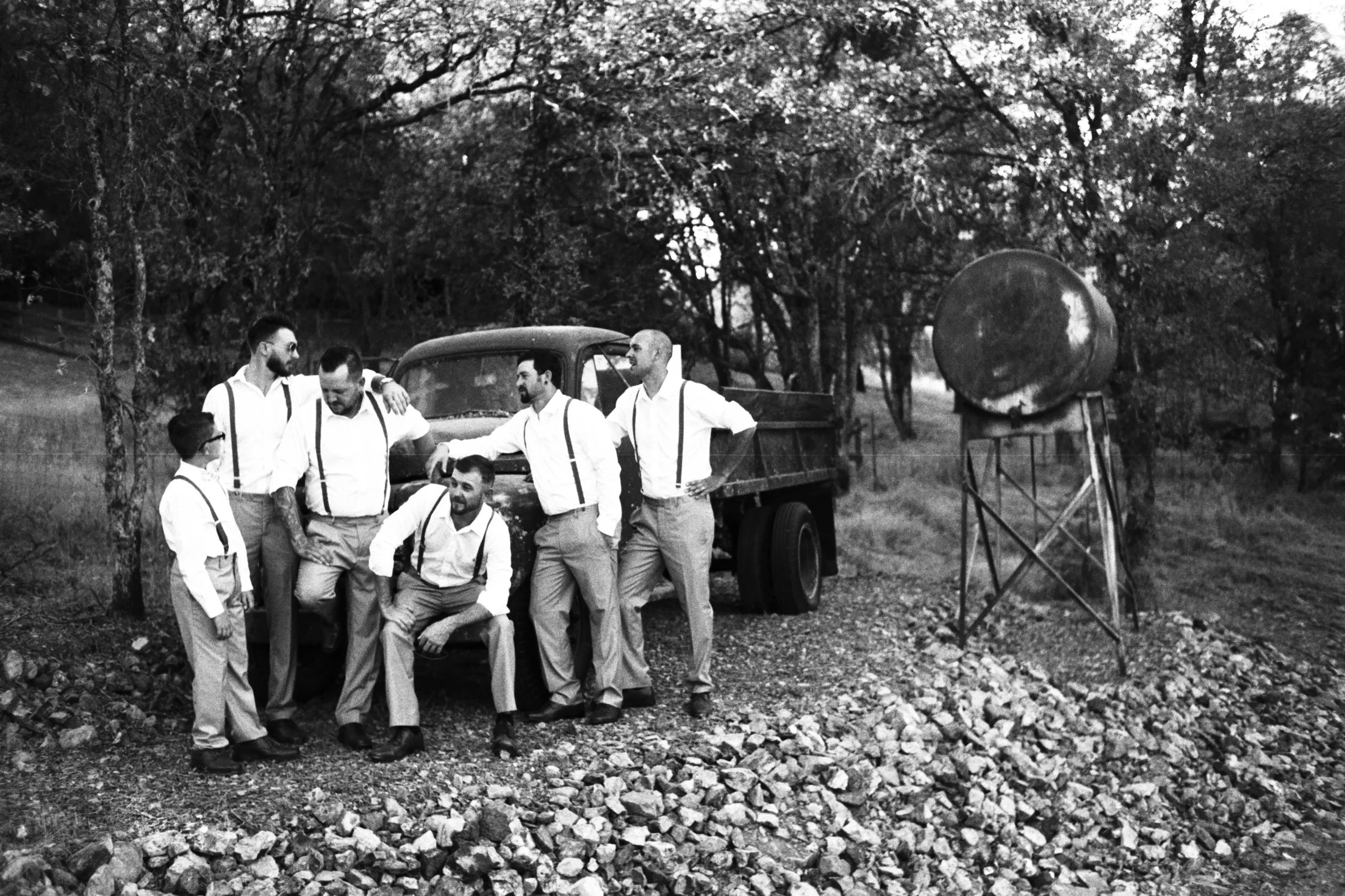 A group of seven men with light skin, dressed in vintage style clothing, pose near an old truck in a wooded outdoor setting with rocks on the ground and a large spherical object on a stand to the right.