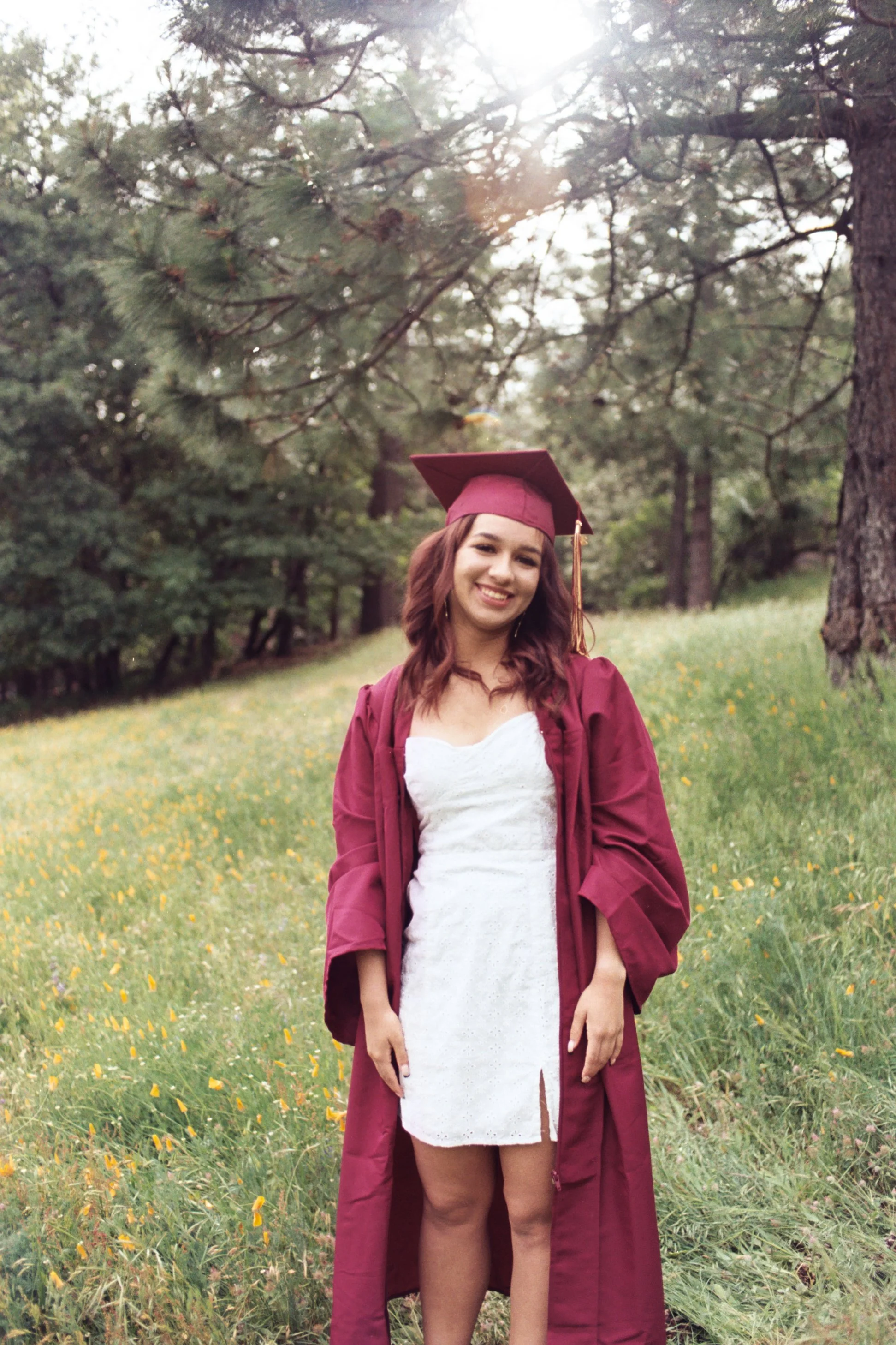 A young woman in a white dress and maroon graduation gown and cap stands outdoors in a grassy, wooded area during daytime with sunlight shining through the trees.