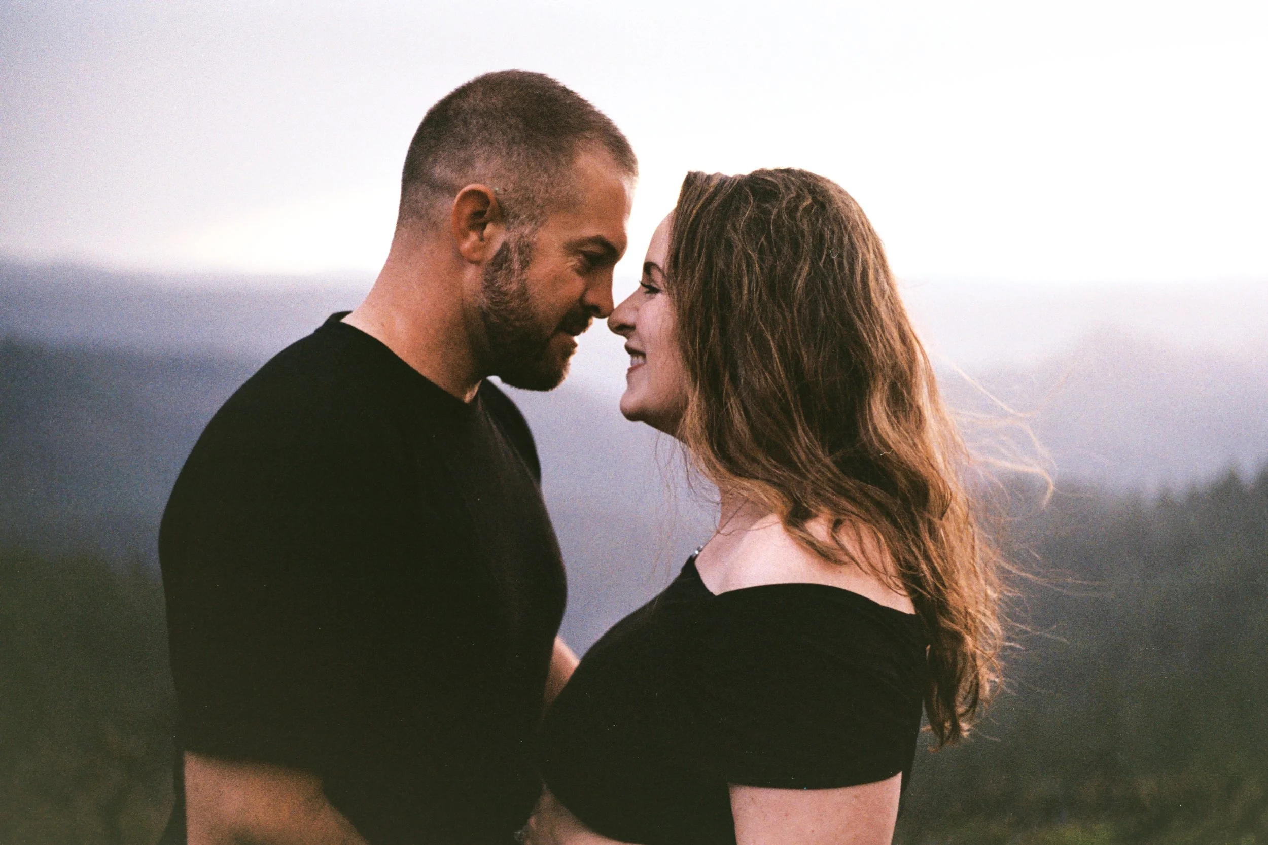 A man and woman with foreheads touching, smiling, outdoors during dusk or dawn.