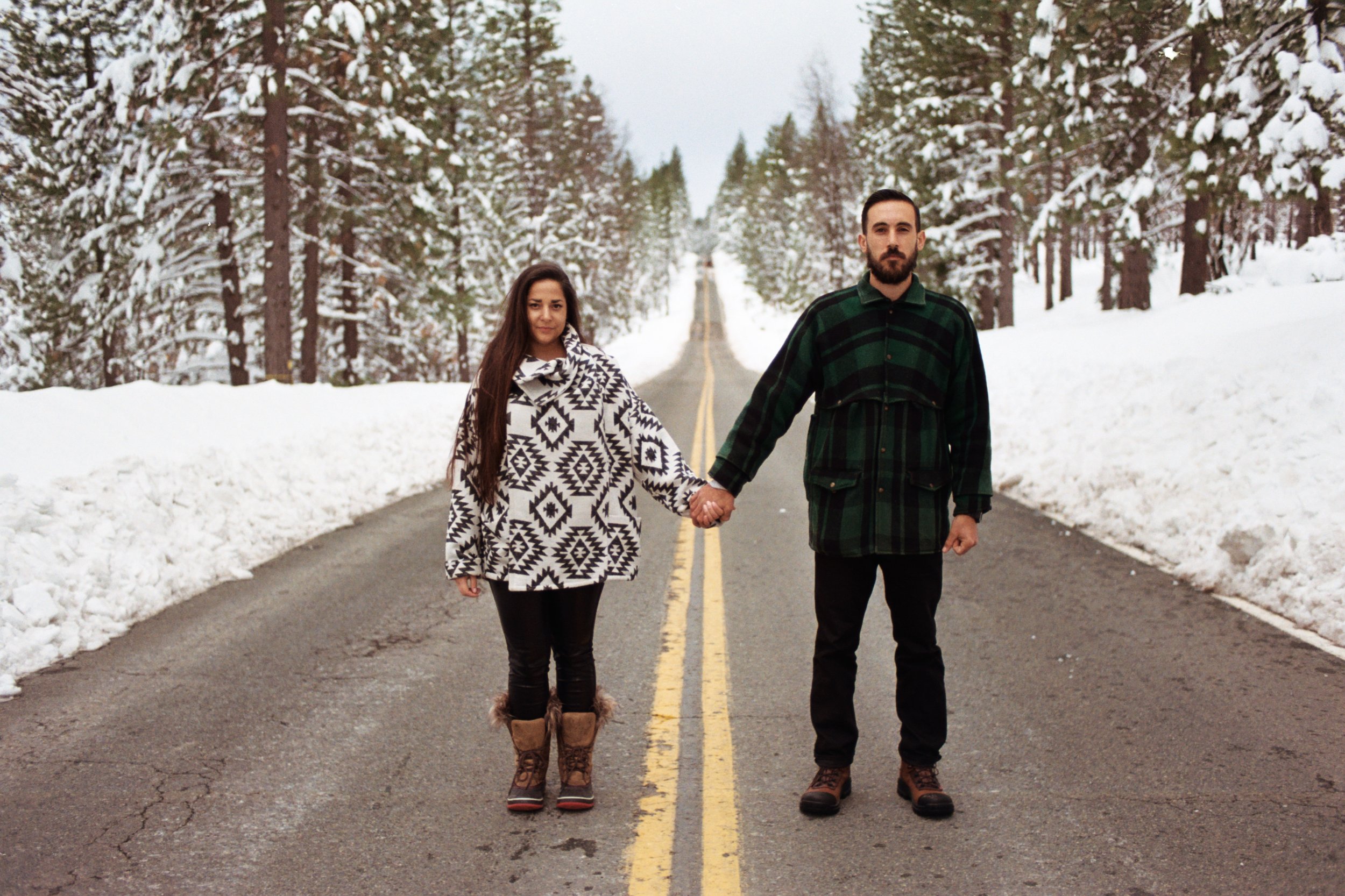 A man and woman holding hands standing on a snow-covered road surrounded by snow-laden tall pine trees in winter.
