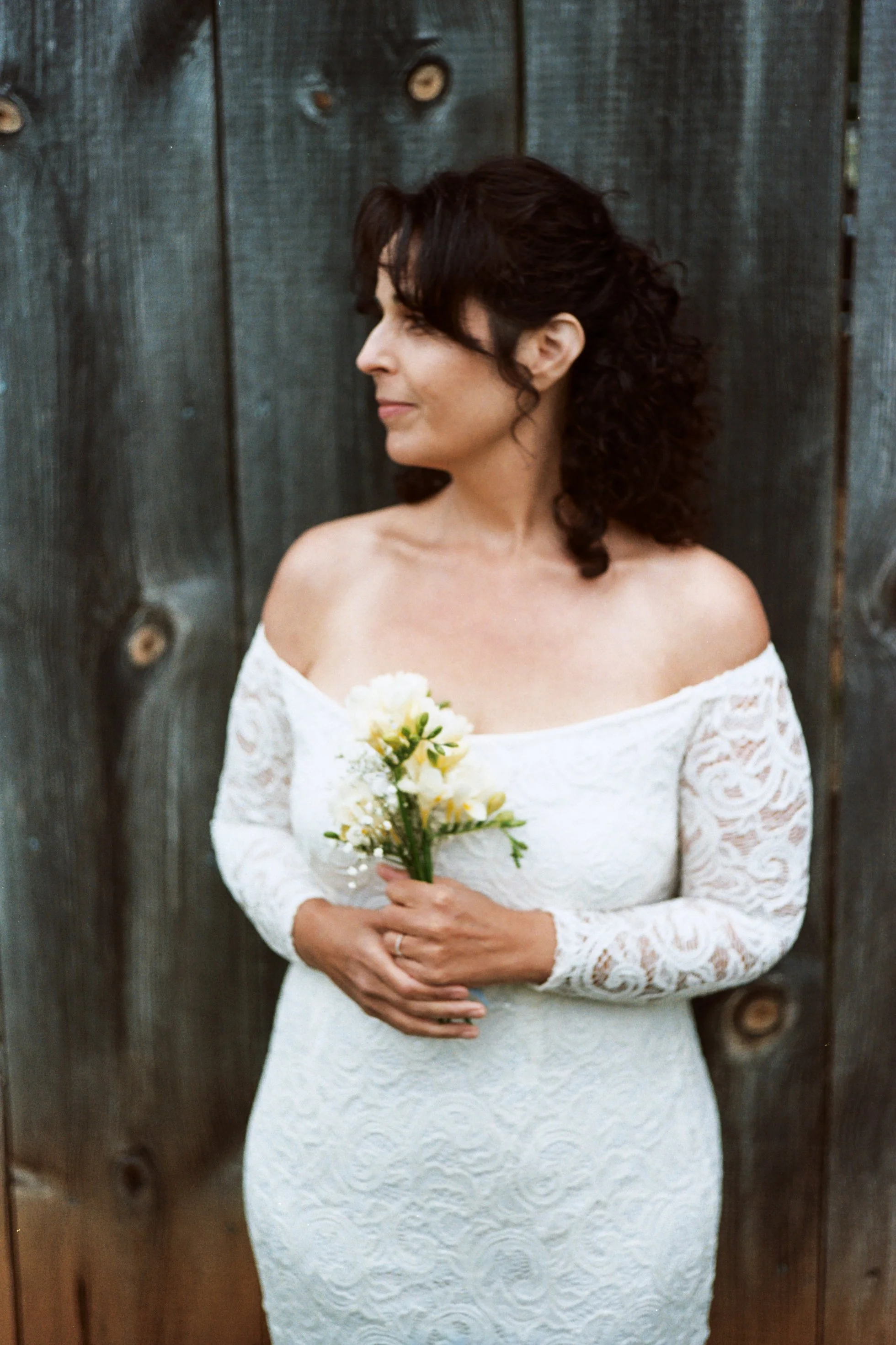 A woman in a white lace off-shoulder dress holding a small bouquet of white and yellow flowers, standing in front of a wooden wall.