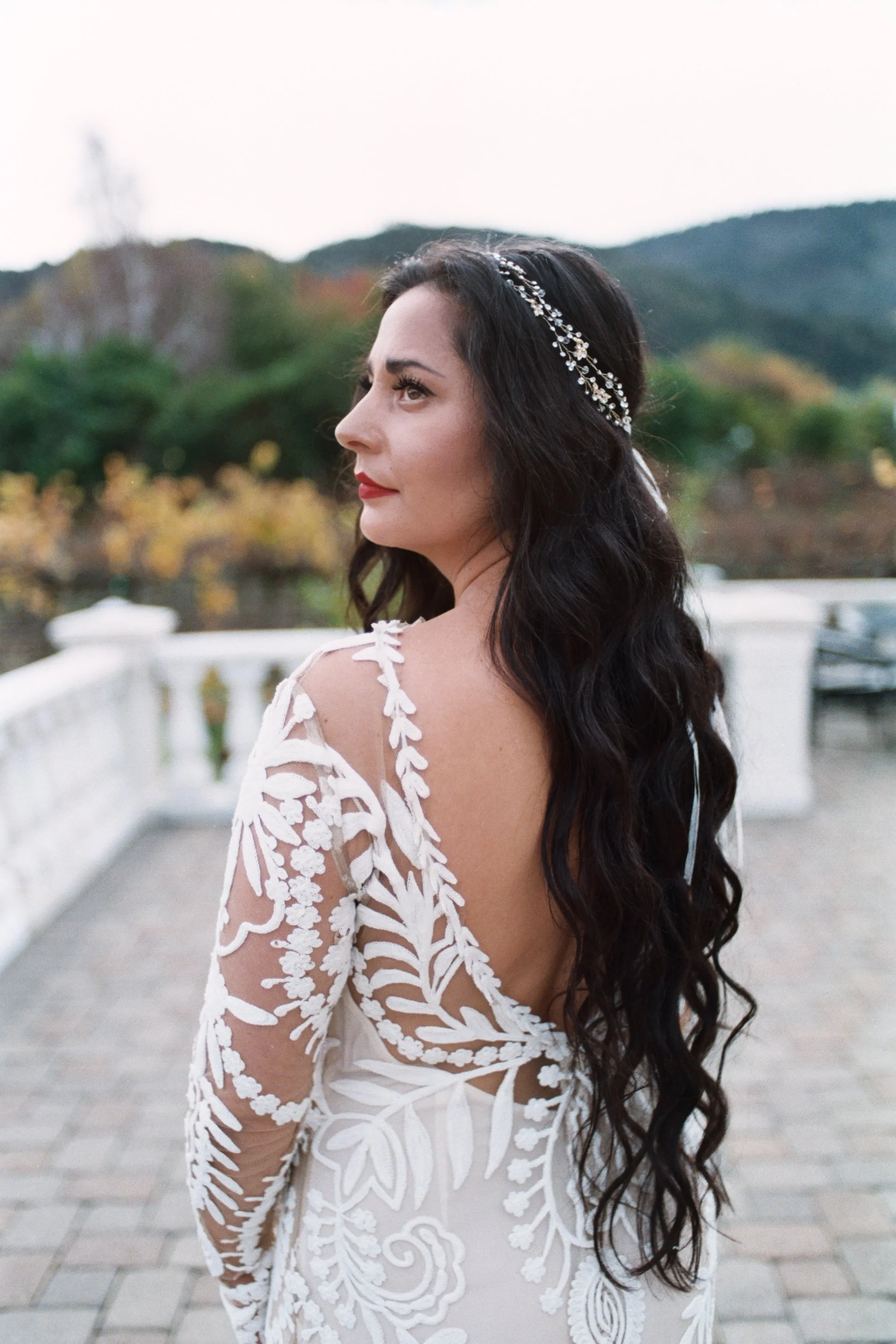 A woman with long dark curly hair and a silver headband, wearing a white dress with floral embroidery, standing outdoors on a bridge with a scenic background of trees and mountains.