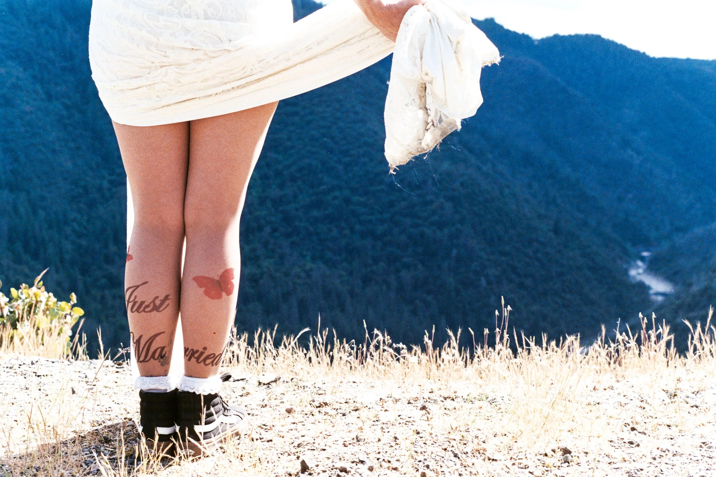 A person's legs with tattoos standing on dry grass, wearing white socks, black shoes, and a white dress with lace, outdoors with mountains in the background.