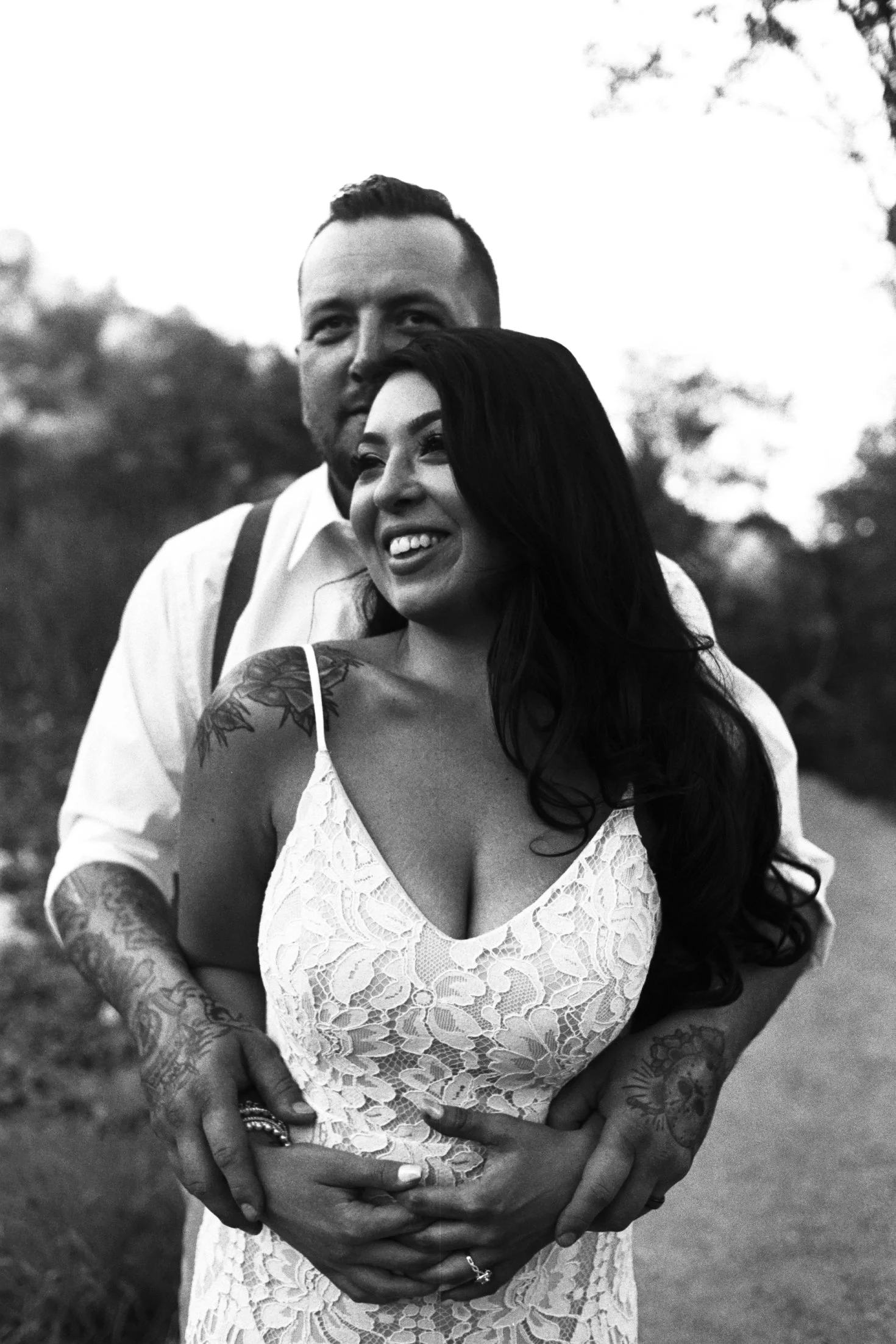 A smiling woman with long dark hair and tattoos on her shoulders and arms wearing a white lace dress, being embraced from behind by a man with short hair and facial hair, dressed in a white shirt and suspenders, outdoors with blurred trees in the bac